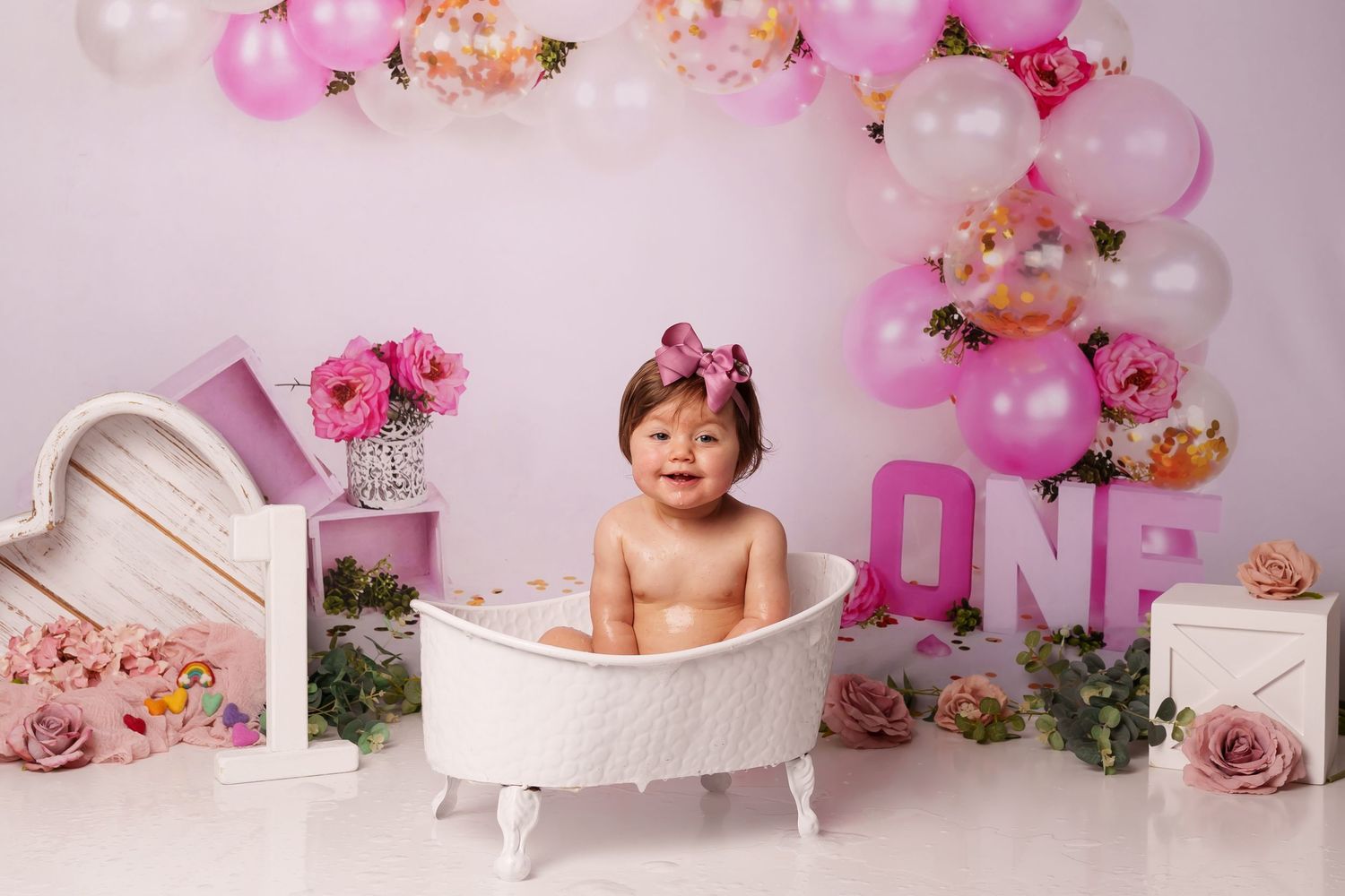 First birthday photo setup with white clawfoot tub, pink balloons, and floral decorations.