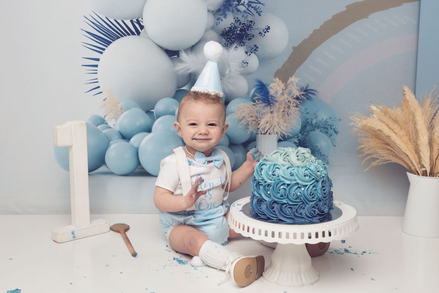 A baby in white outfit sits next to a blue cake with balloon decorations for a first birthday shoot.