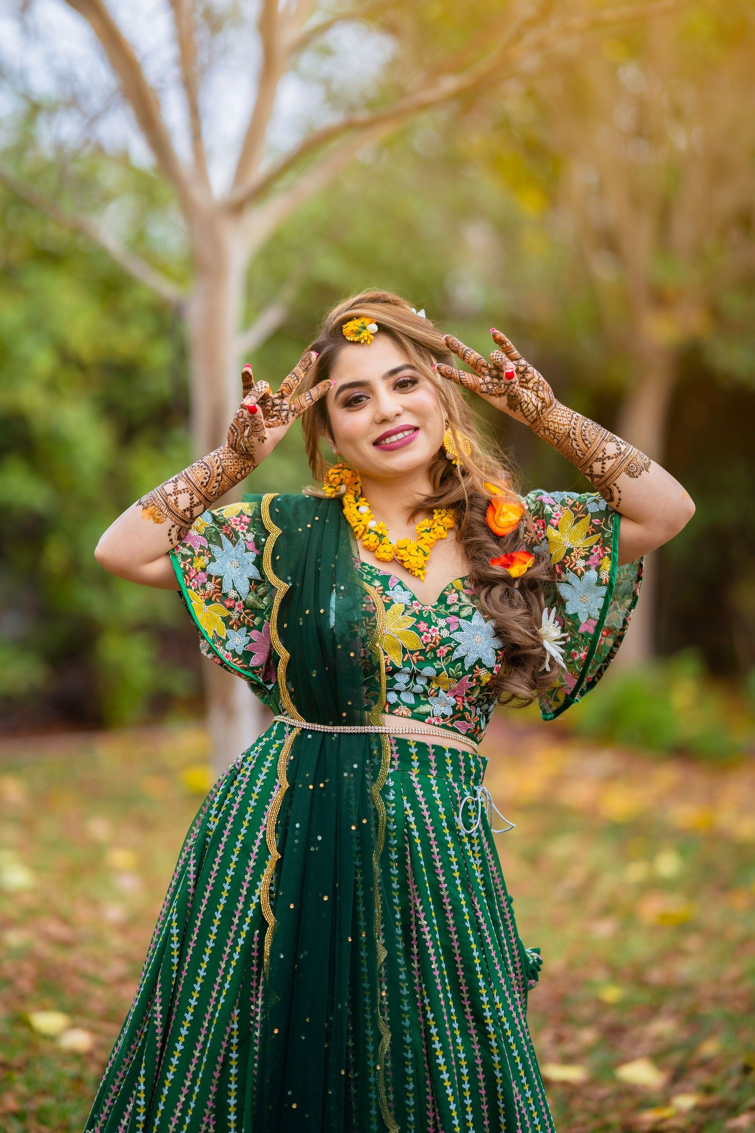 Traditional Indian bride in emerald green lehenga with intricate henna designs poses playfully in autumn garden setting.