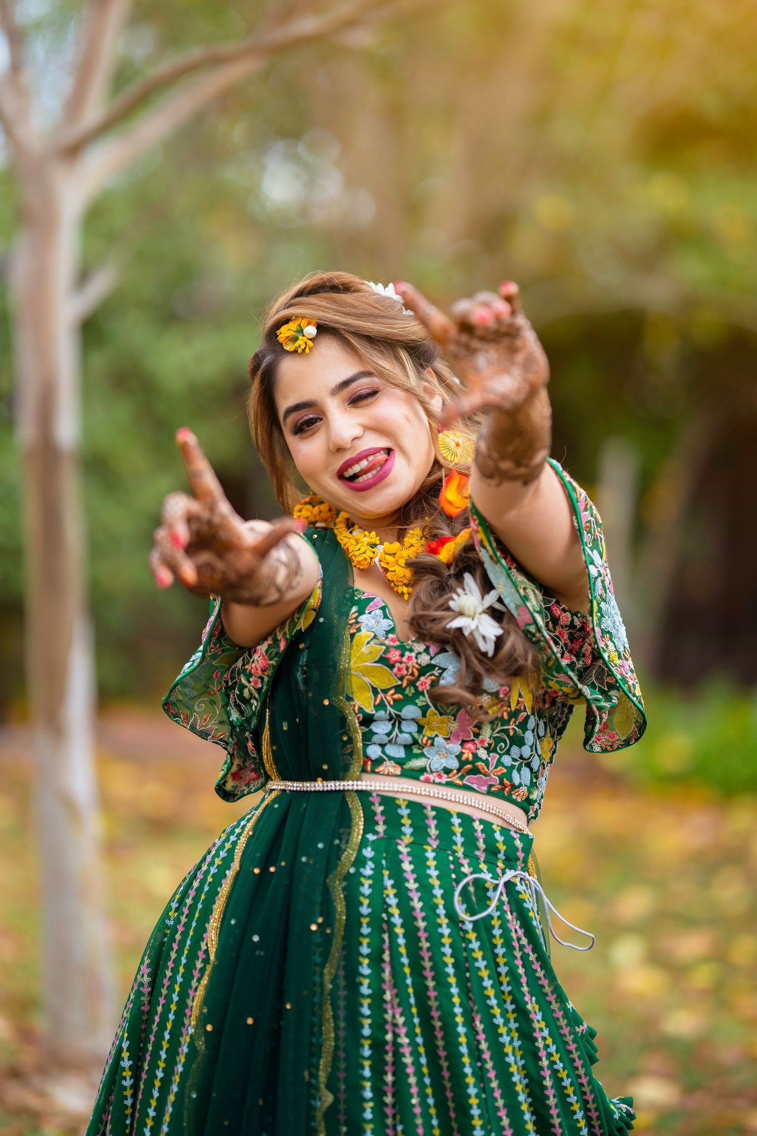 Joyful pose in embellished green lehenga with ornate henna designs against autumn foliage.