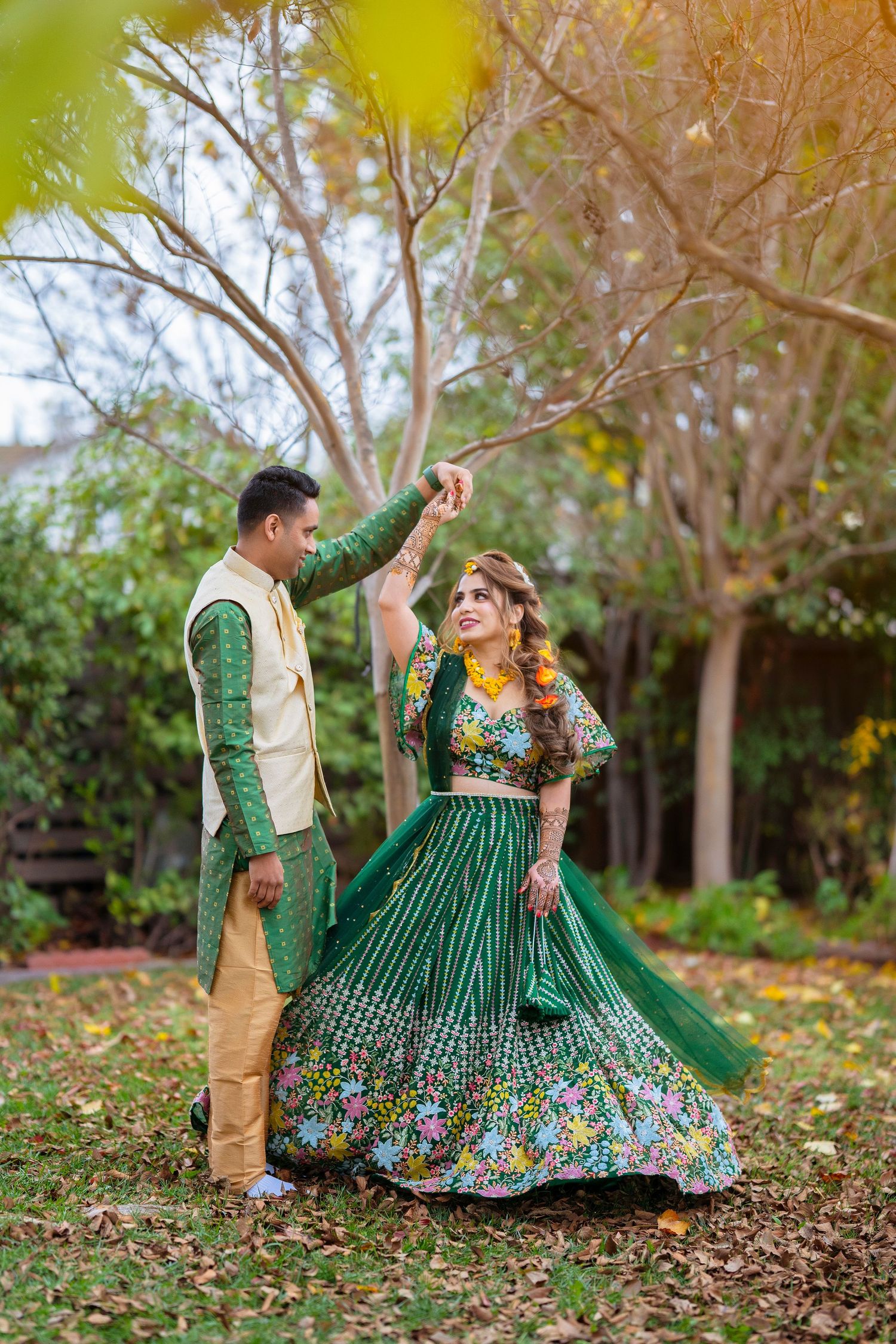 Couple in coordinating green and cream Indian attire dance together among fall leaves.