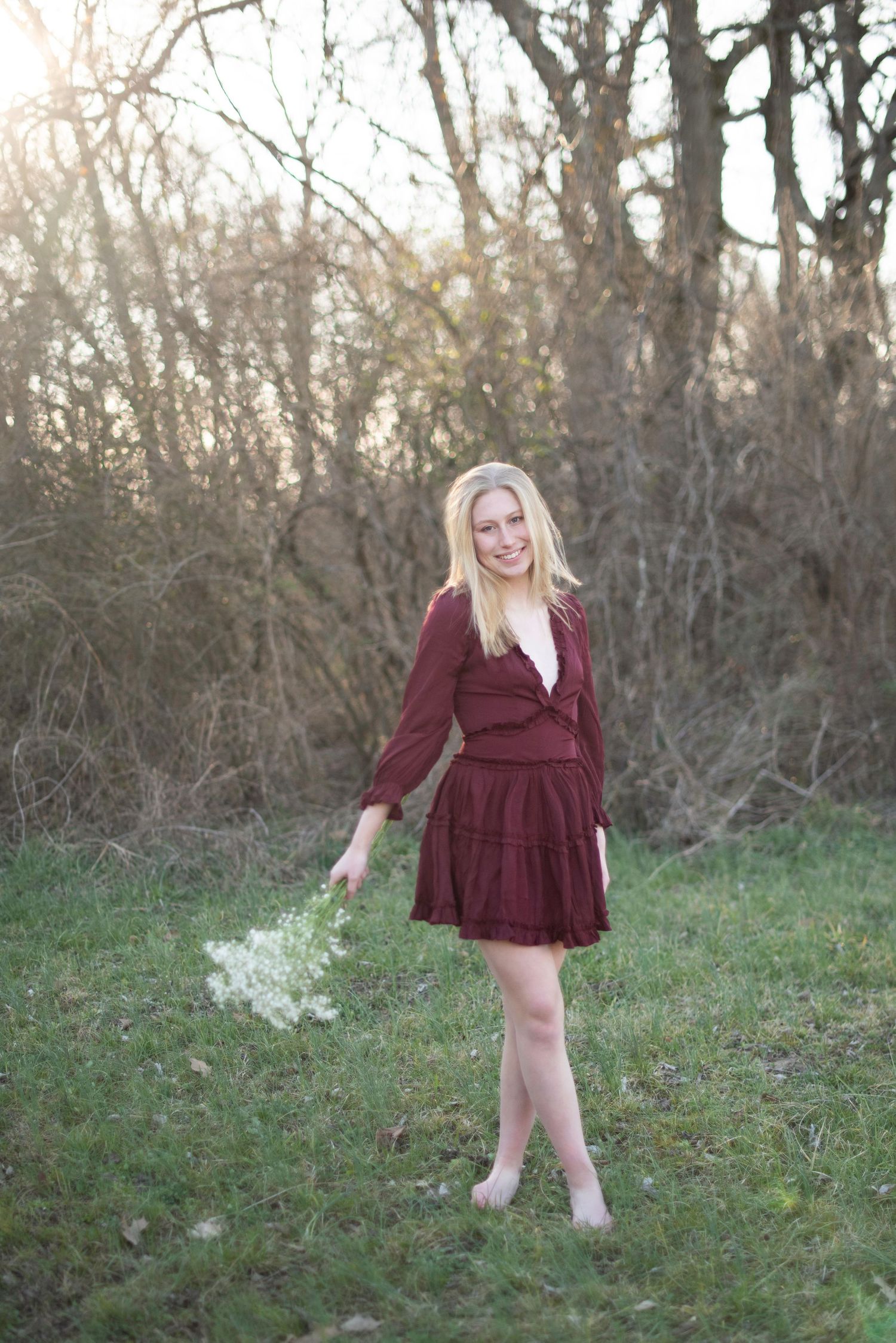 Playful pose in burgundy dress standing on grass with sunlit trees in background.