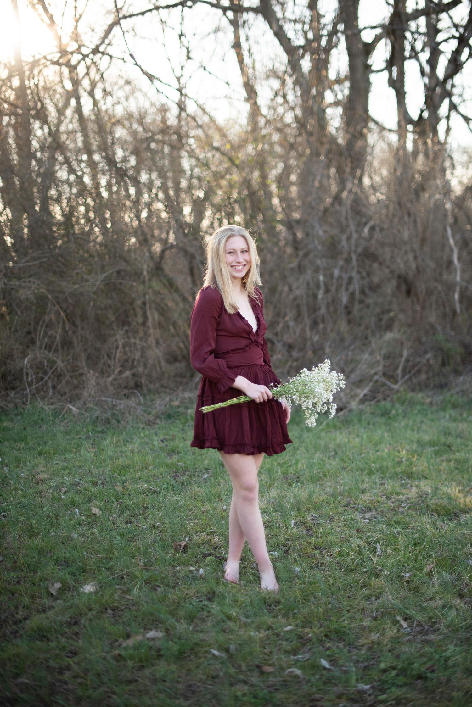 A figure in a burgundy dress holds white flowers while standing in a grassy outdoor setting at dusk.