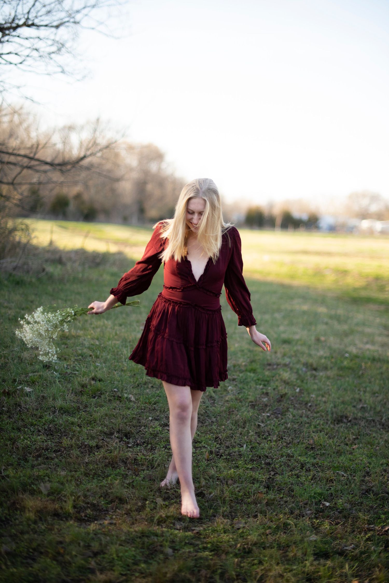 Person in flowing burgundy dress walking barefoot through grass at sunset.