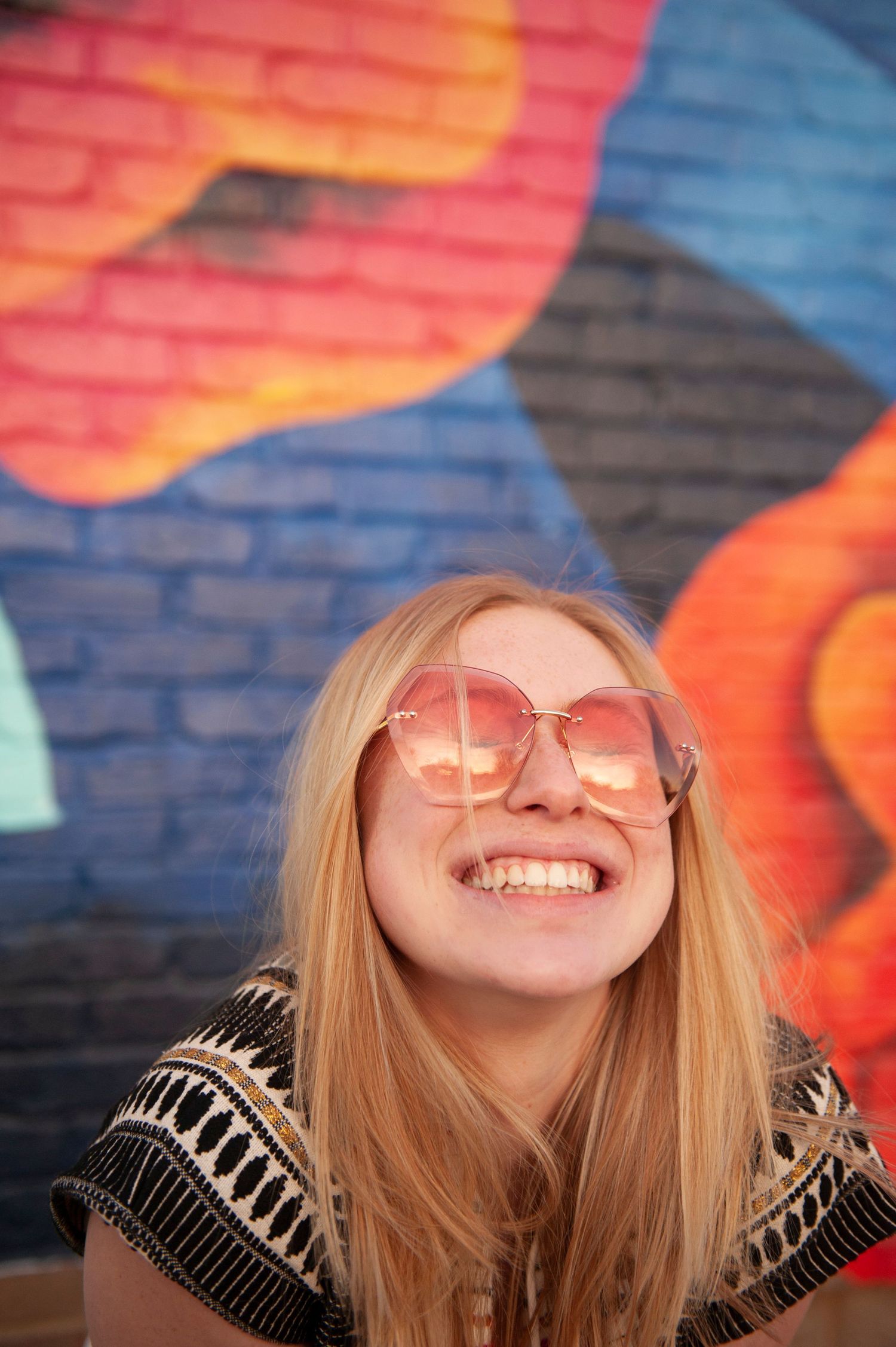 Woman with blonde hair wearing pink sunglasses smiles in front of colorful street art mural.