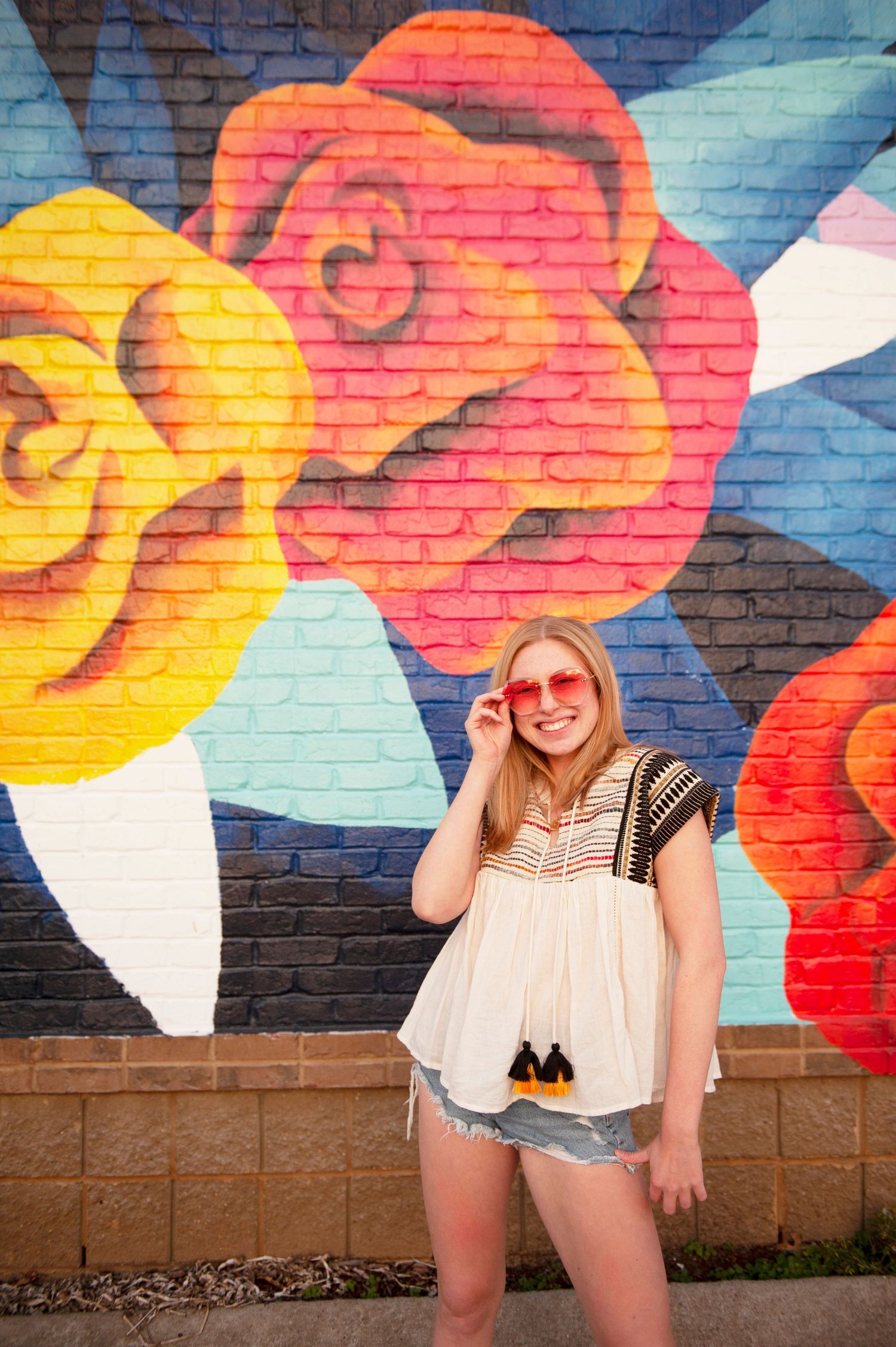 Standing by colorful rose mural wearing boho style top and denim shorts.