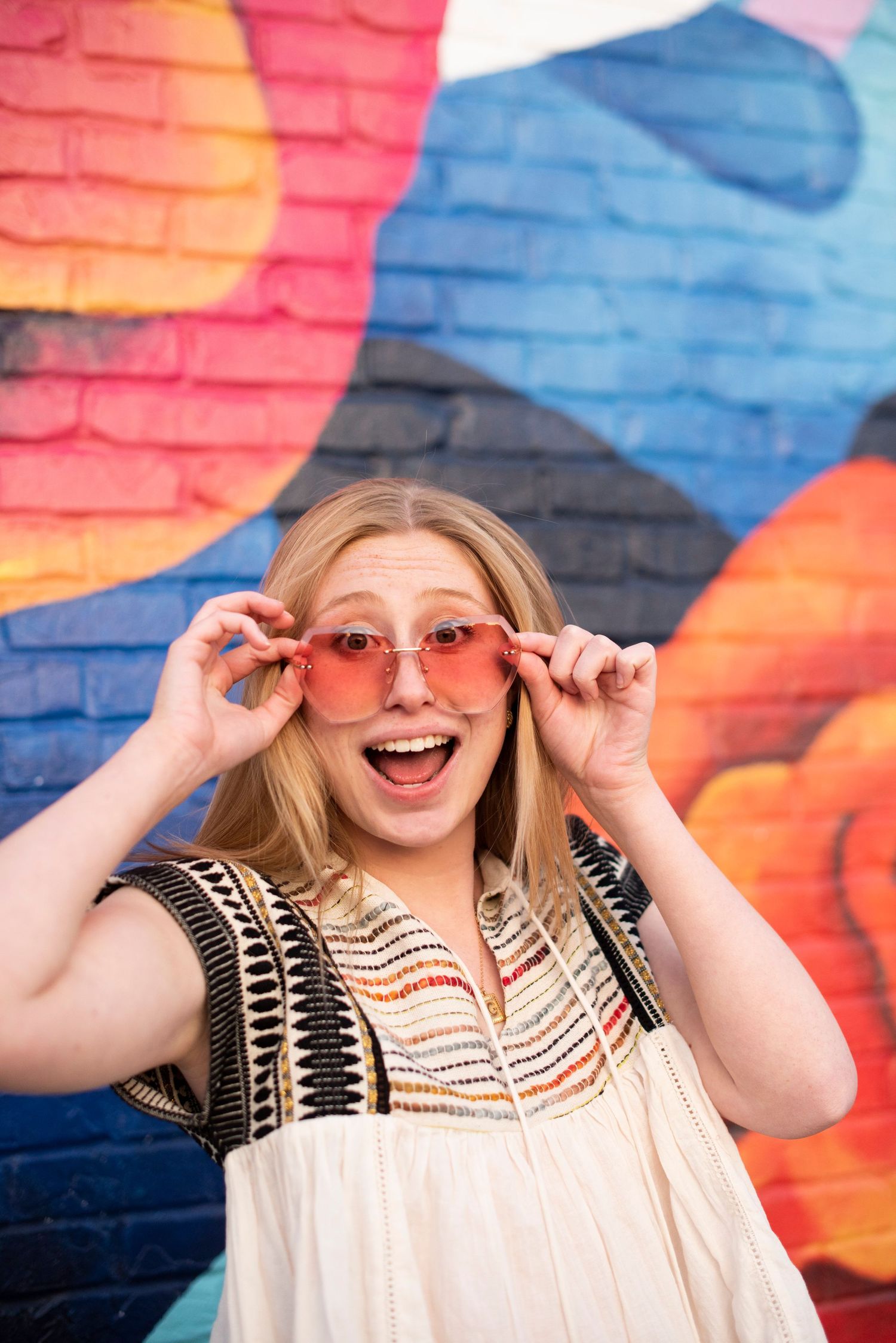 Playful pose adjusting pink sunglasses with excited expression against painted wall.