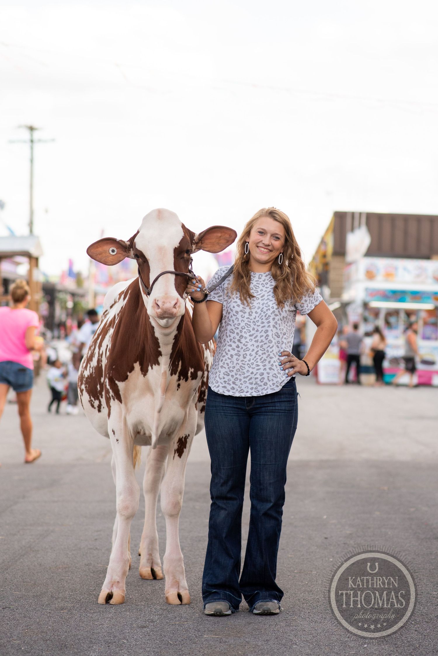 A brown and white dairy cow stands next to someone at a county fair on a sunny day.