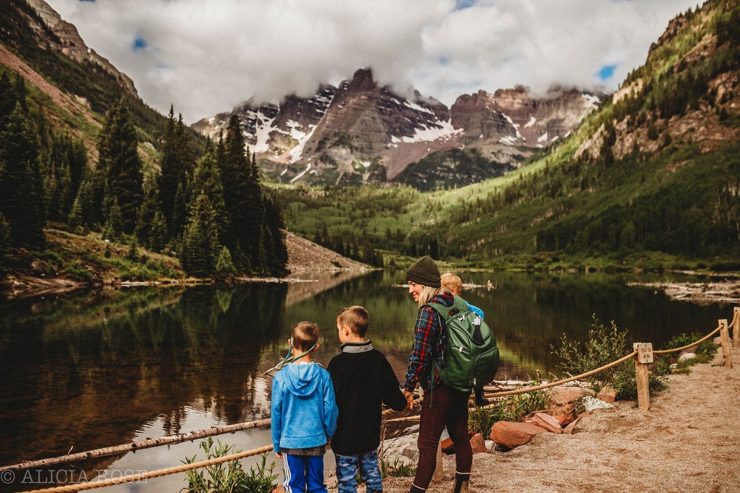 Three people admire a scenic mountain lake view with dramatic peaks in the background.