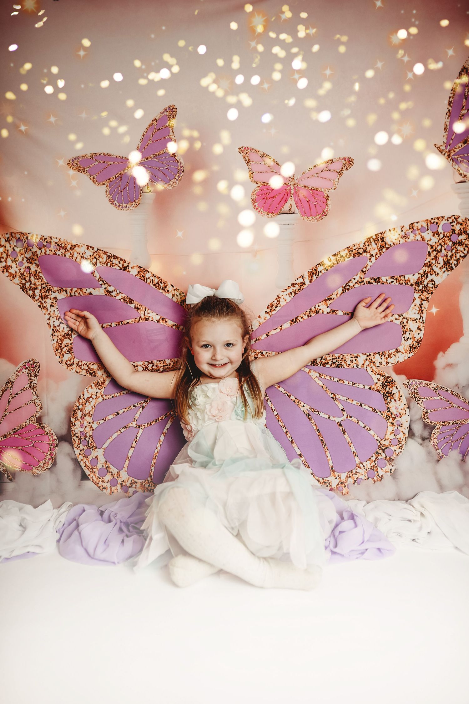 Child in white dress posing with sparkly purple butterfly wings against glittery backdrop.
