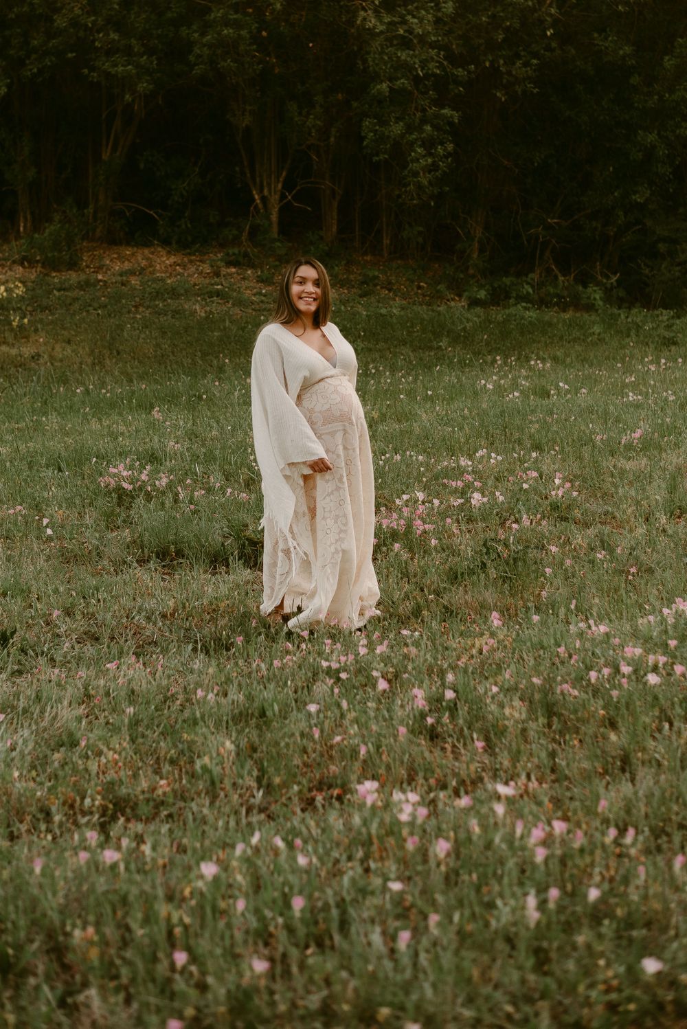 Maternity portrait in lace dress amid blooming spring meadow with forest backdrop.