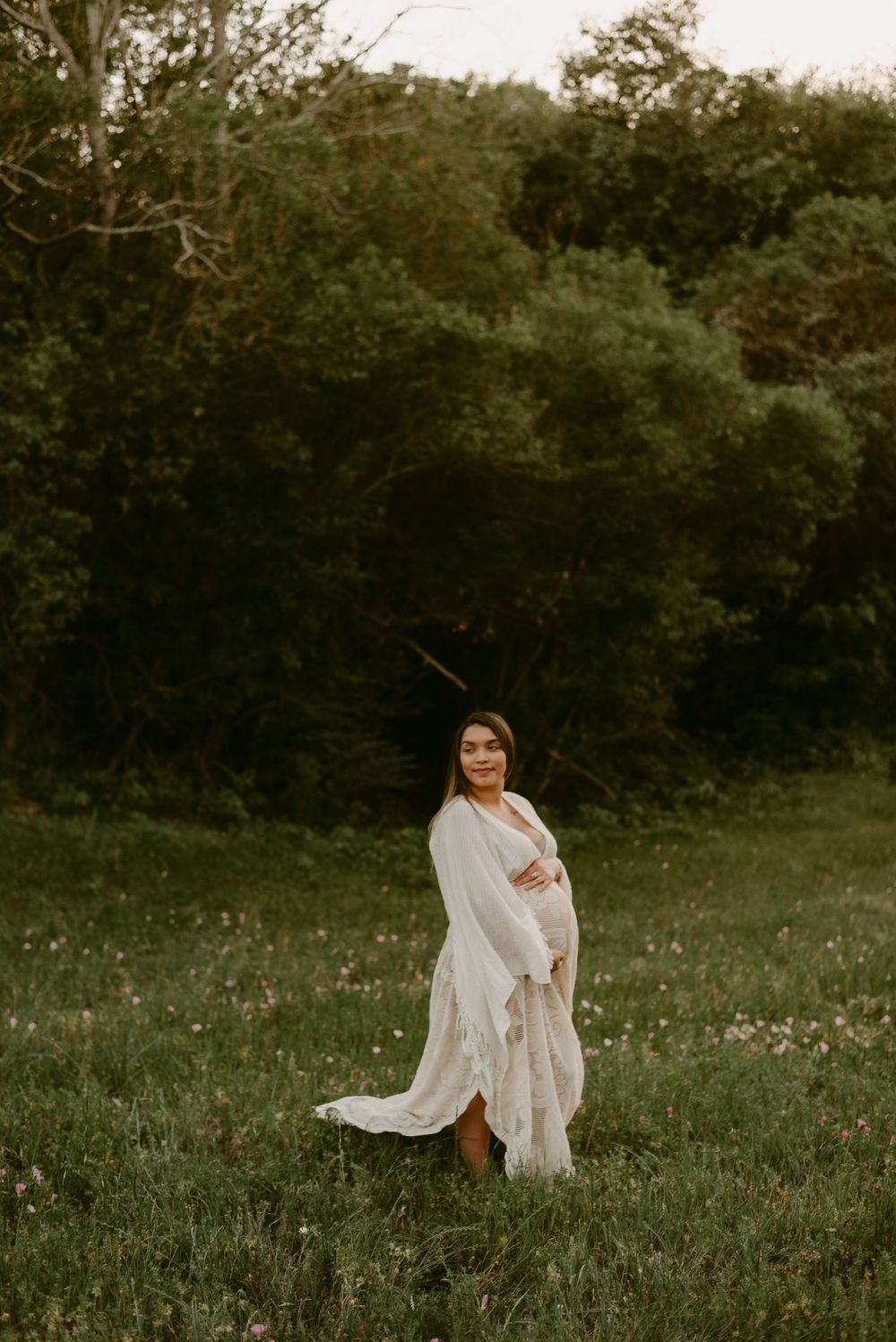 Ethereal maternity portrait of woman in flowing white dress standing in sunlit meadow.