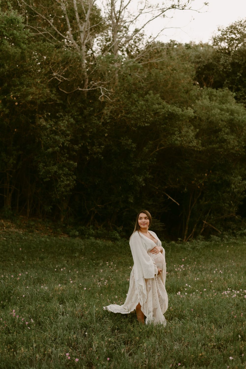 Ethereal maternity photo in white dress standing in a wildflower meadow at golden hour.