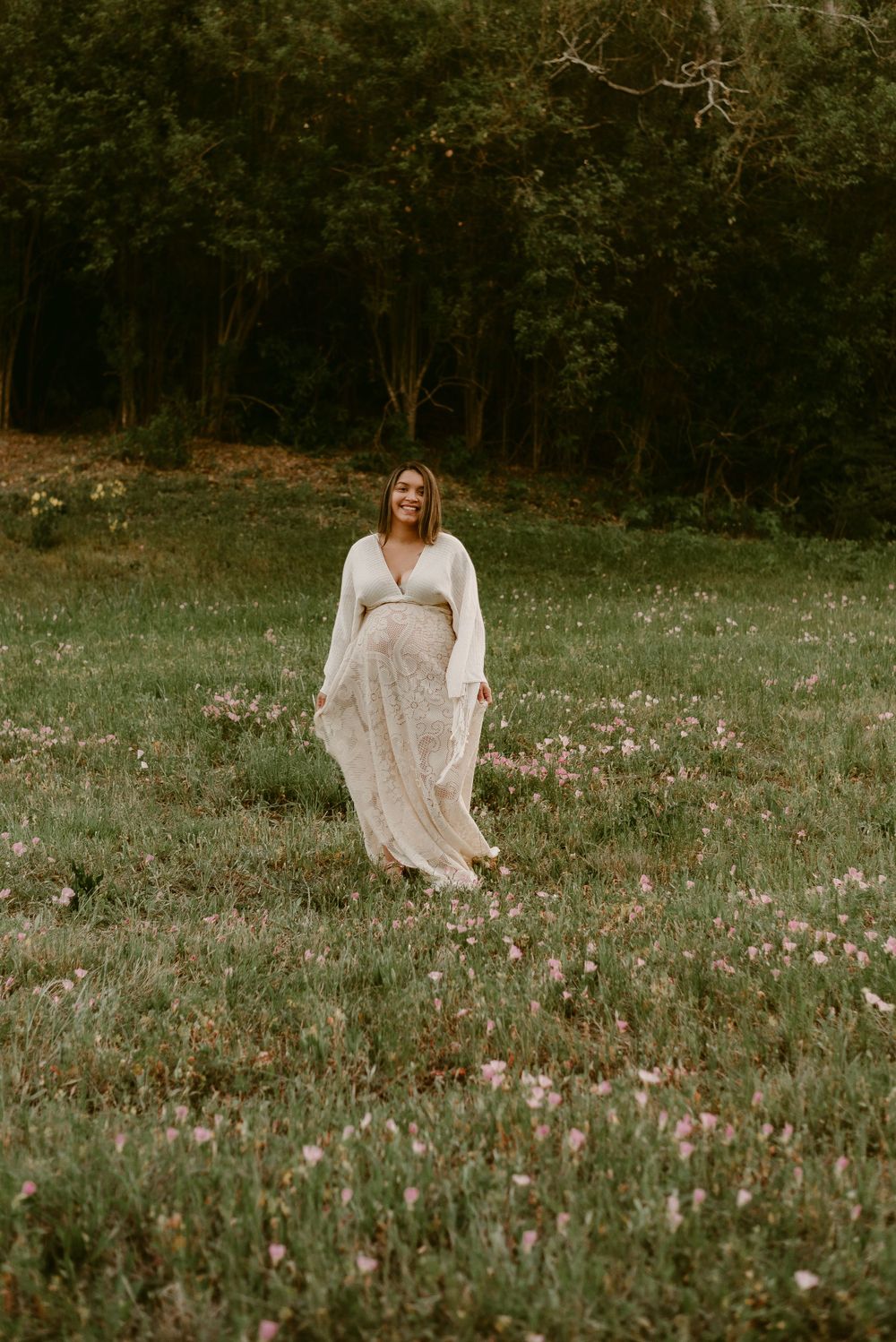Expecting mother in flowing white dress poses in field of wildflowers at dusk.