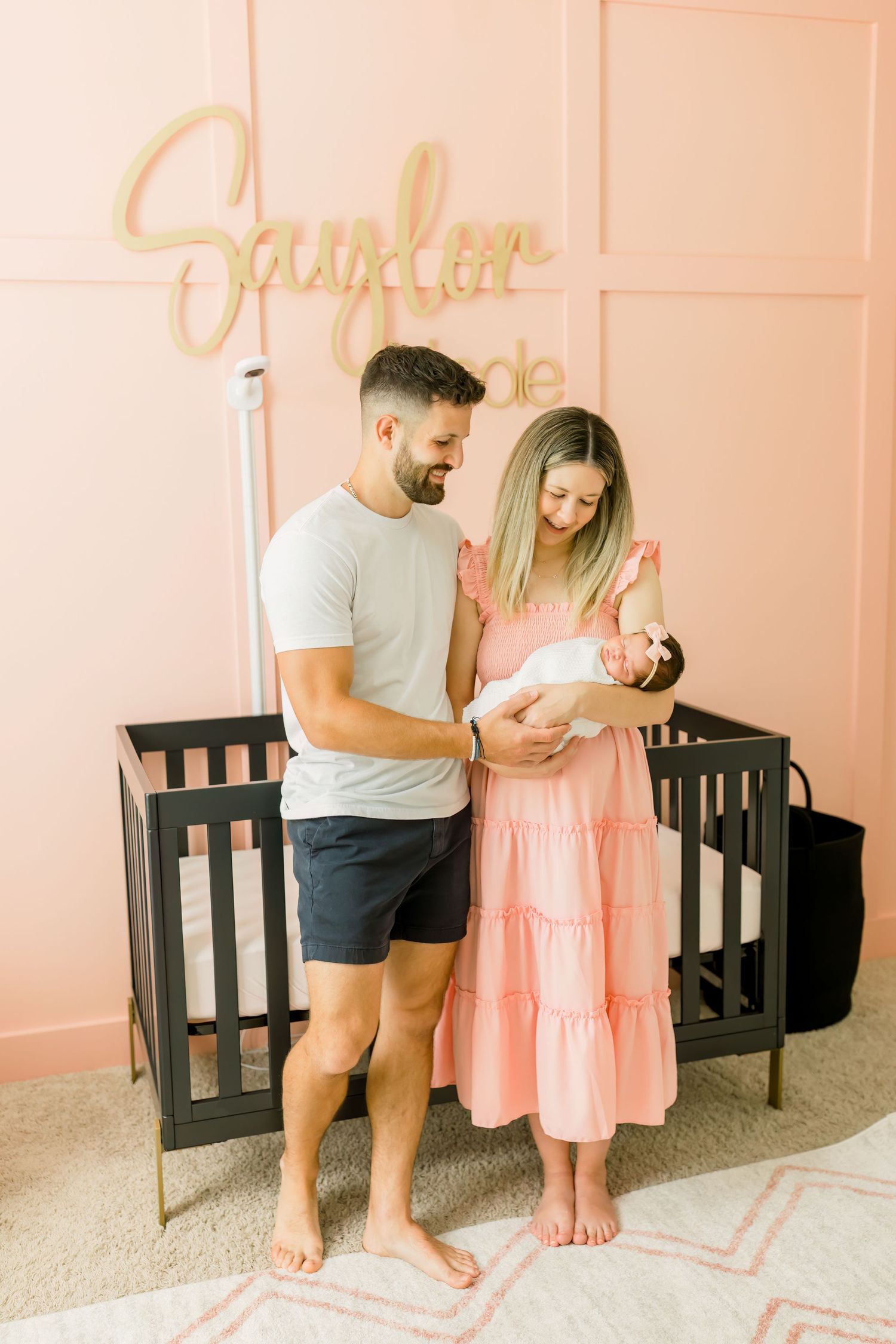 Parents standing with their newborn in front of a pink wall with decorative gold lettering.