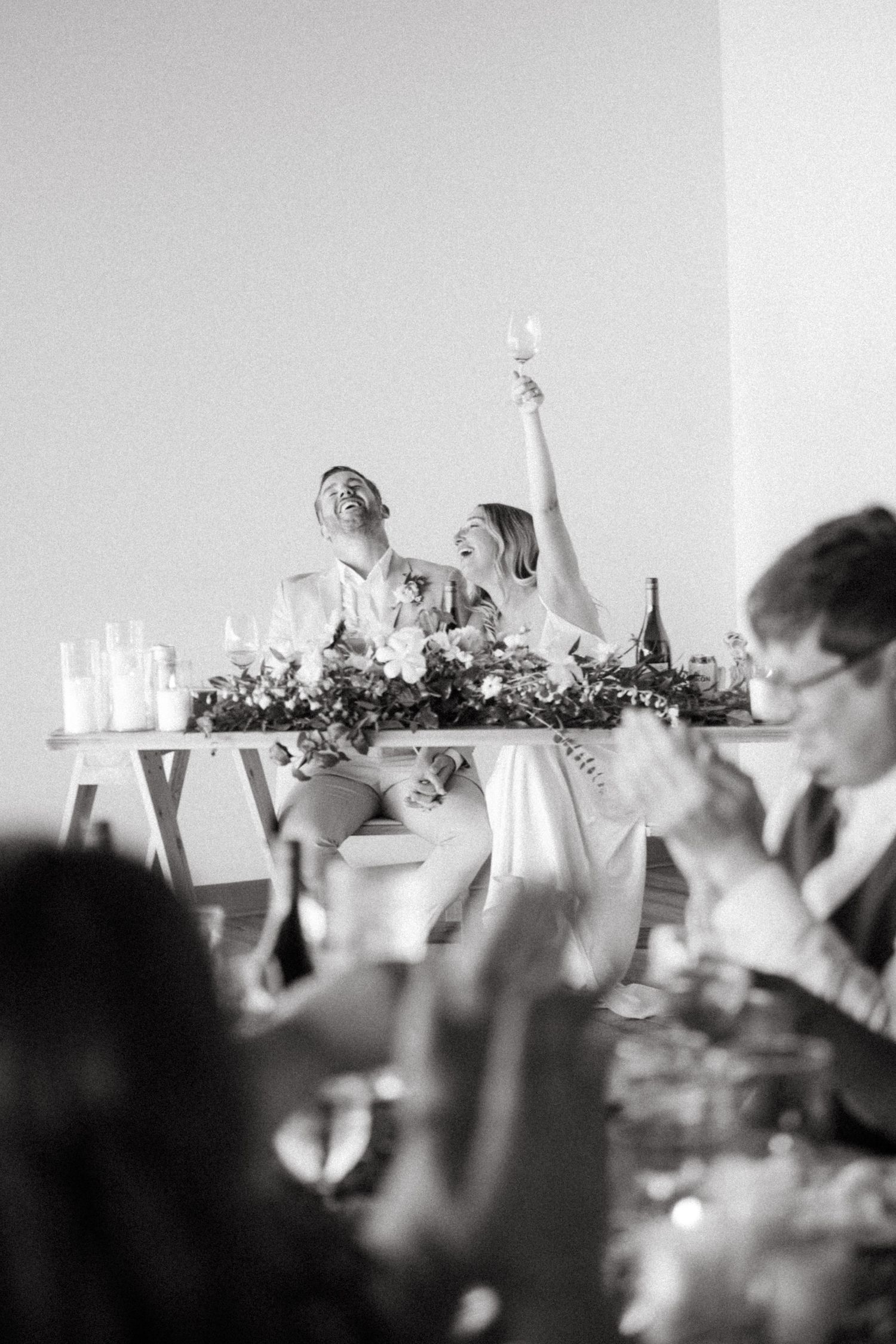 Black and white photo of joyful newlyweds raising glasses during wedding reception toast.