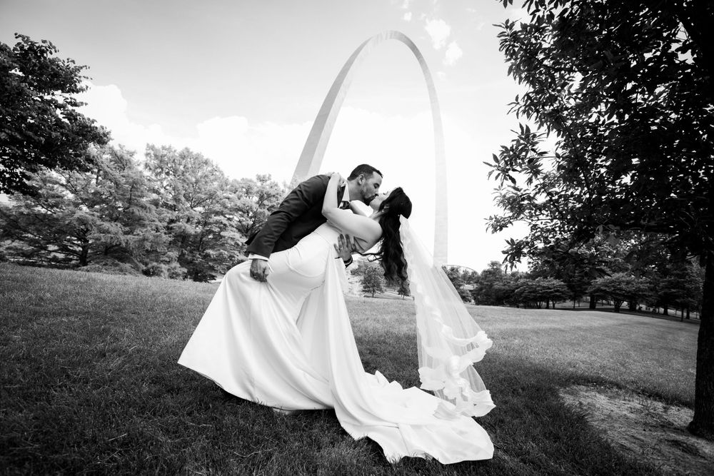 A black and white photo of a groom dipping and kissing his bride in front of the St. Louis Arch