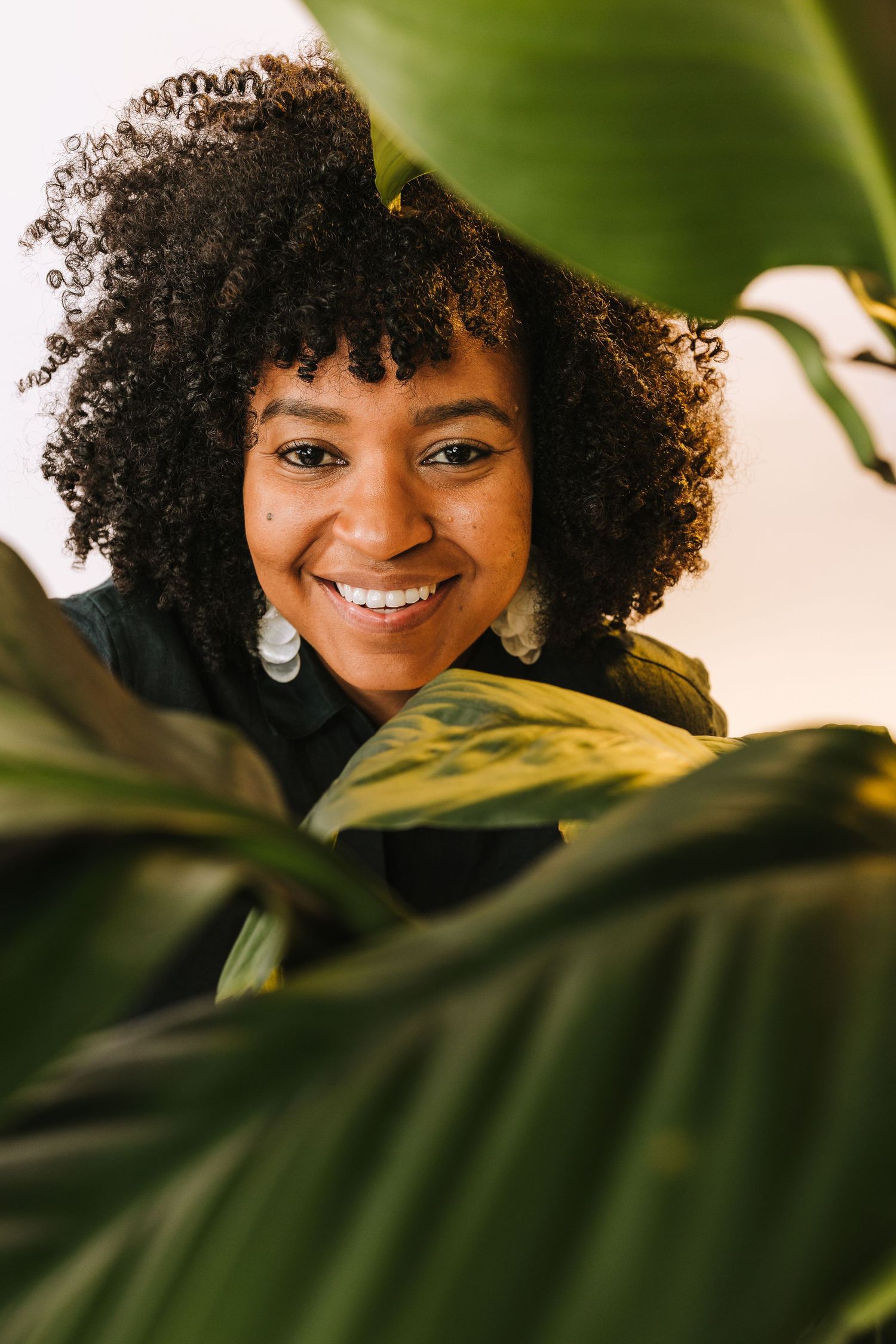 Portrait framed by green tropical leaves showing natural curly hair and silver earrings with a warm smile.