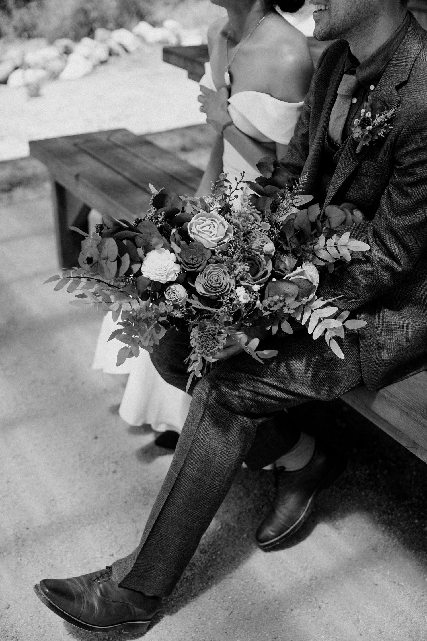 A black and white detail shot of a wedding bouquet with roses and greenery held against formal attire.