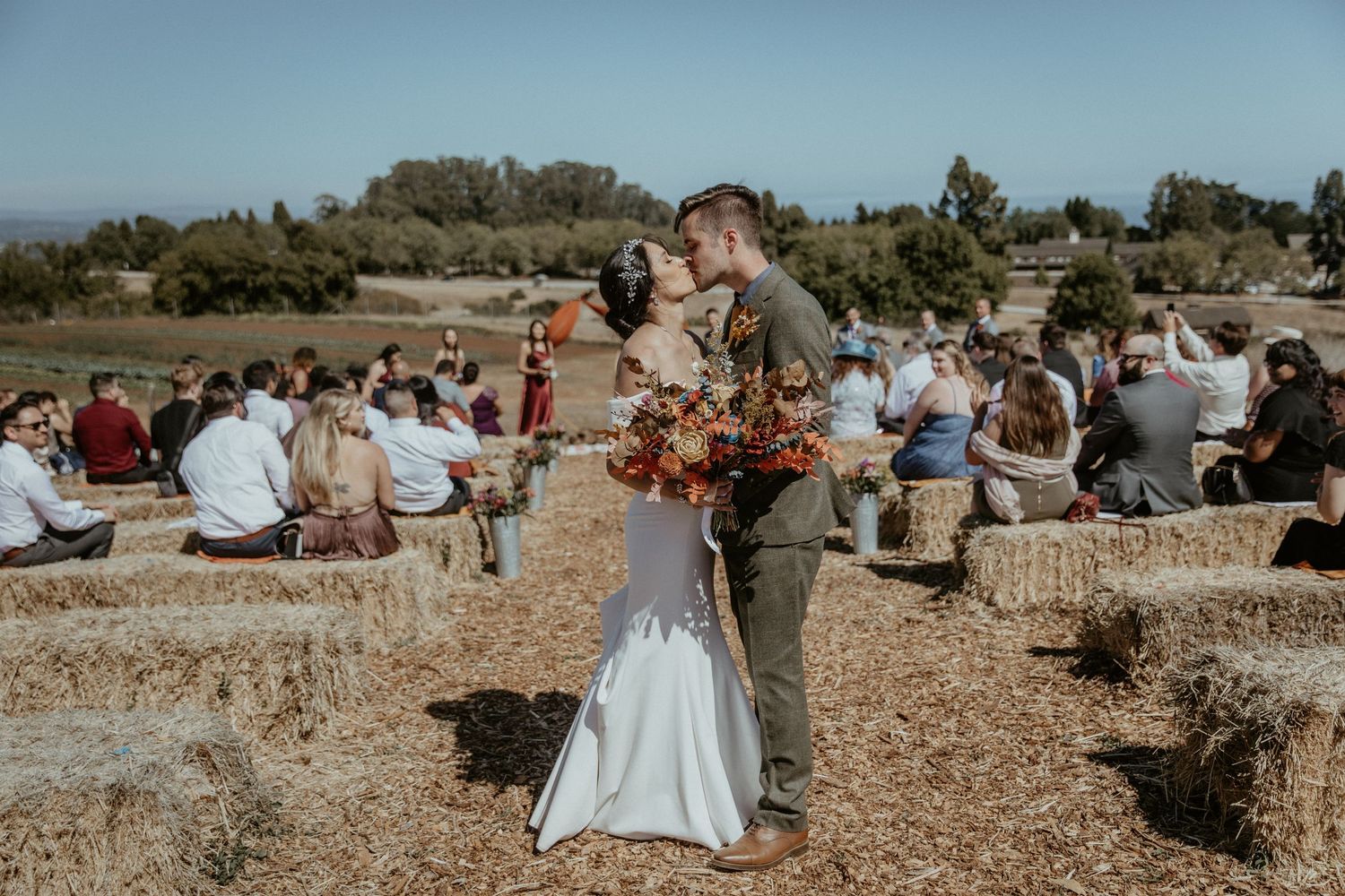 An outdoor wedding ceremony in a rustic setting with hay bale seating and natural scenery in the background.