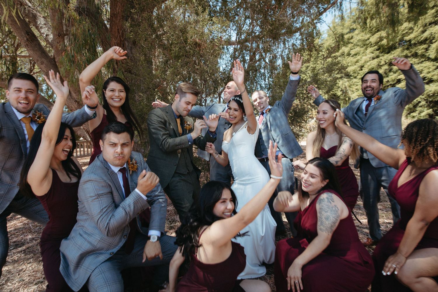 Wedding party celebrates with energetic dance moves outdoors among trees.