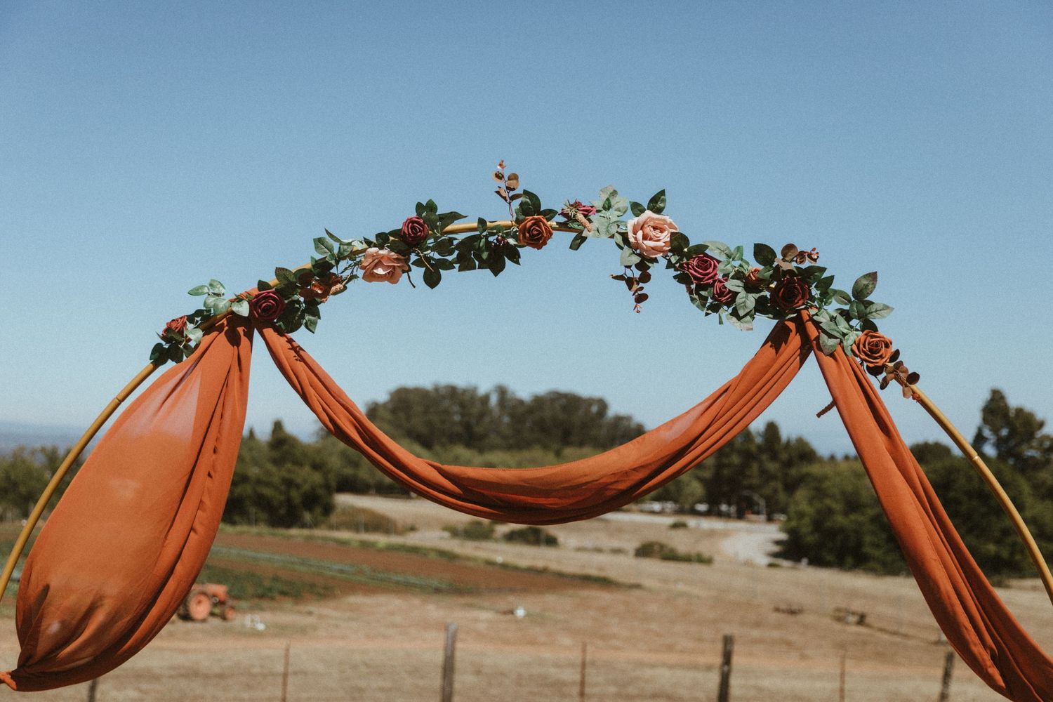 A terracotta-colored fabric arch decorated with greenery and flowers against a blue sky.