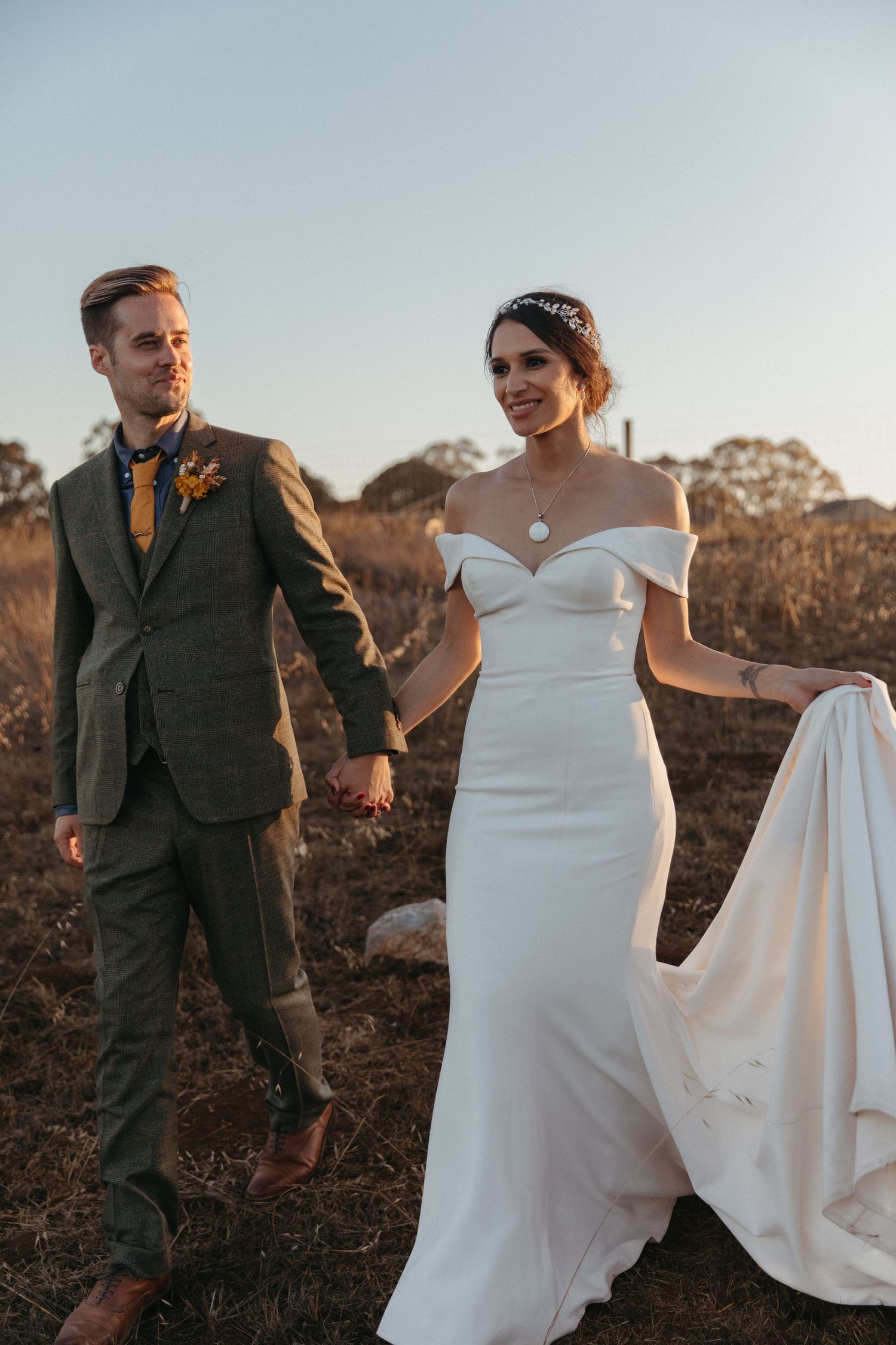 Sunset wedding photo of couple walking hand in hand through field in formal attire.