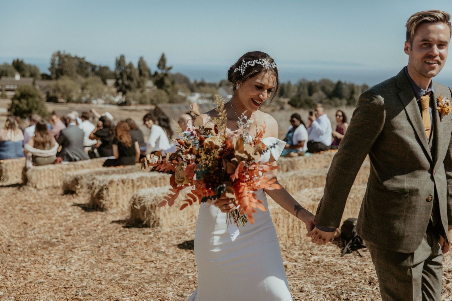 Wedding couple walking in rustic outdoor setting with autumn-colored bouquet.