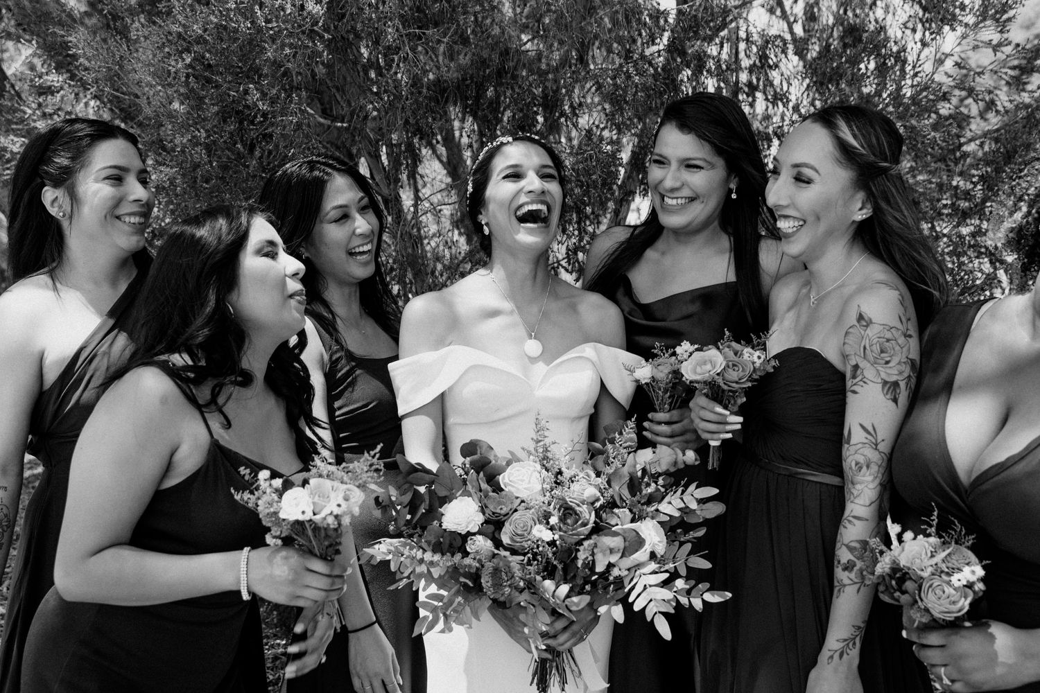 A candid black and white wedding photo showing a bride and bridesmaids in black dresses laughing together outdoors.