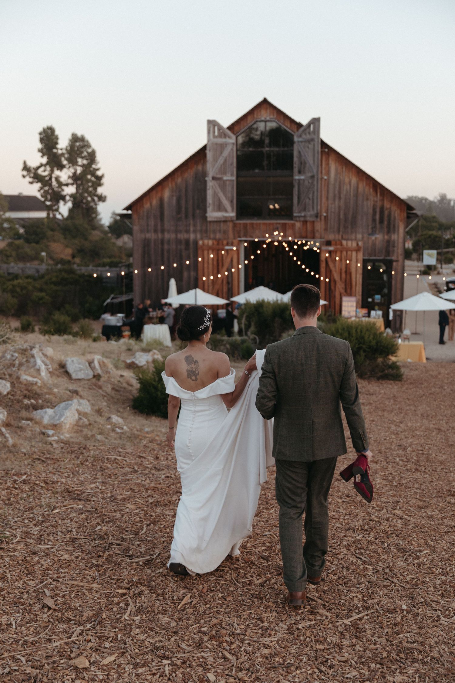 A couple walks towards a rustic barn decorated with string lights at dusk.