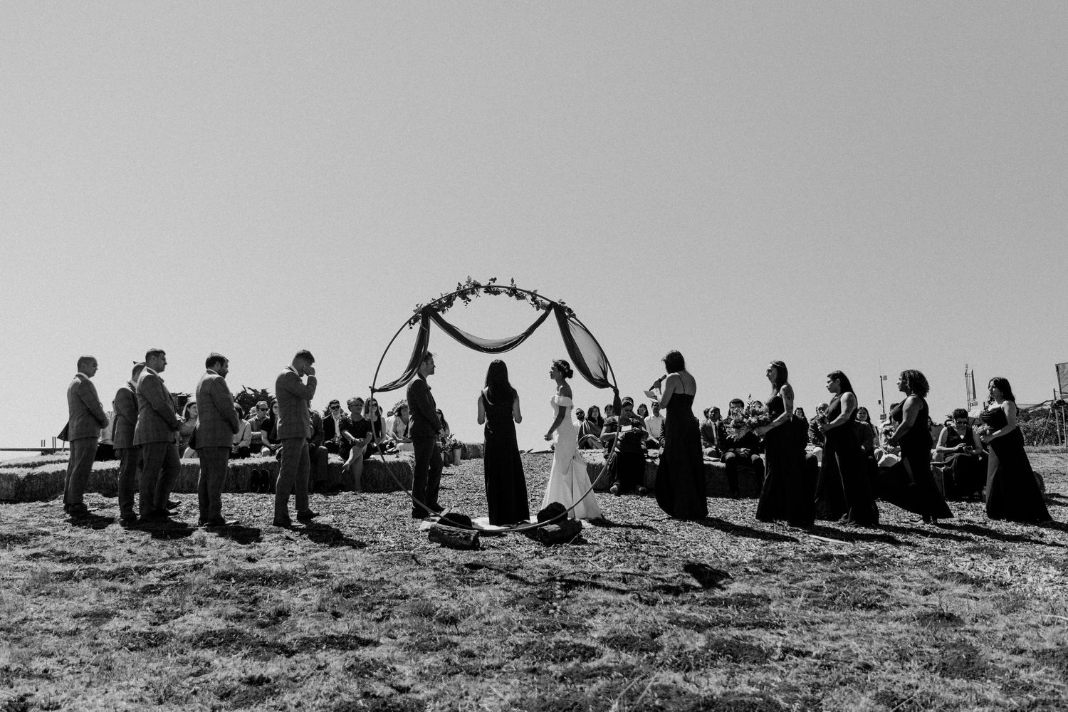 A vintage black and white photograph of an outdoor wedding ceremony with guests gathered in a circle on a hillside.