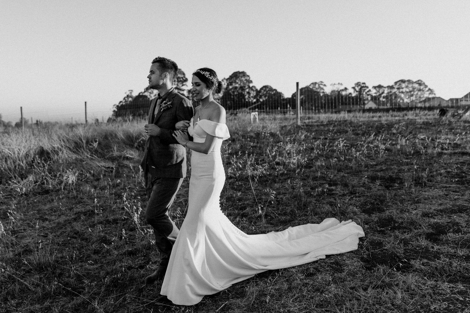 Wedding couple walking together in field with bride's dress trailing behind.