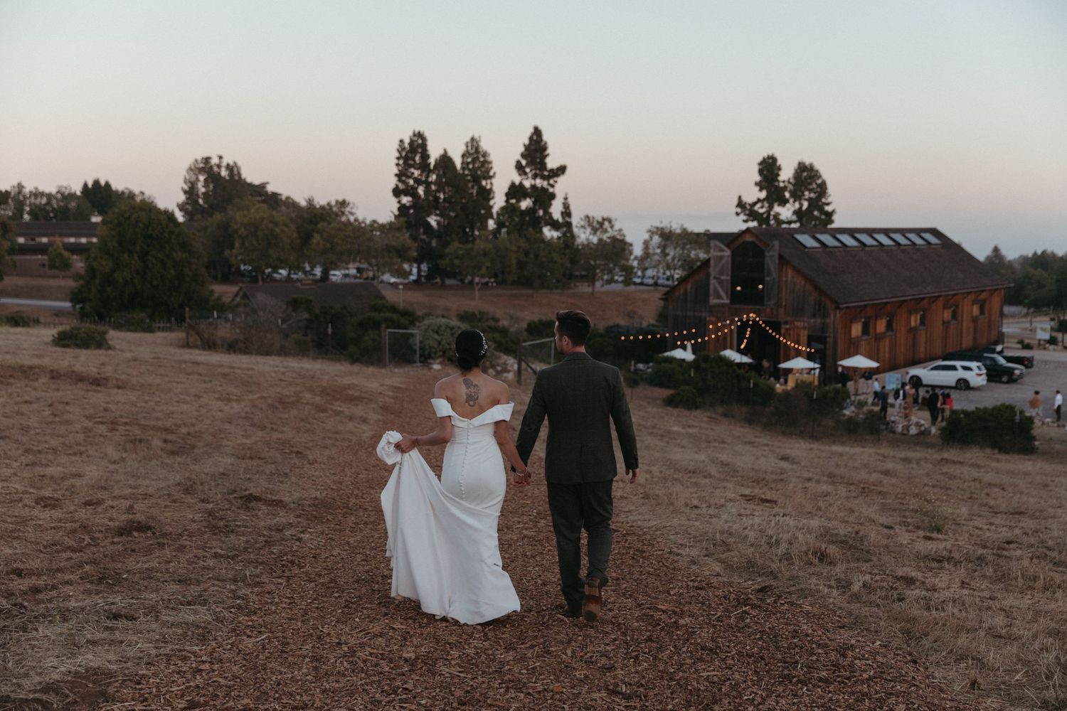 A couple walks hand in hand toward a rustic barn venue at sunset.