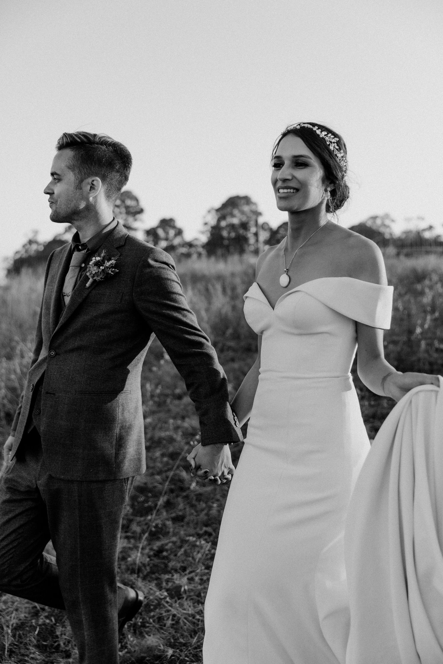 Black and white photo of a bride in off-shoulder dress and groom in suit walking through a field at sunset.