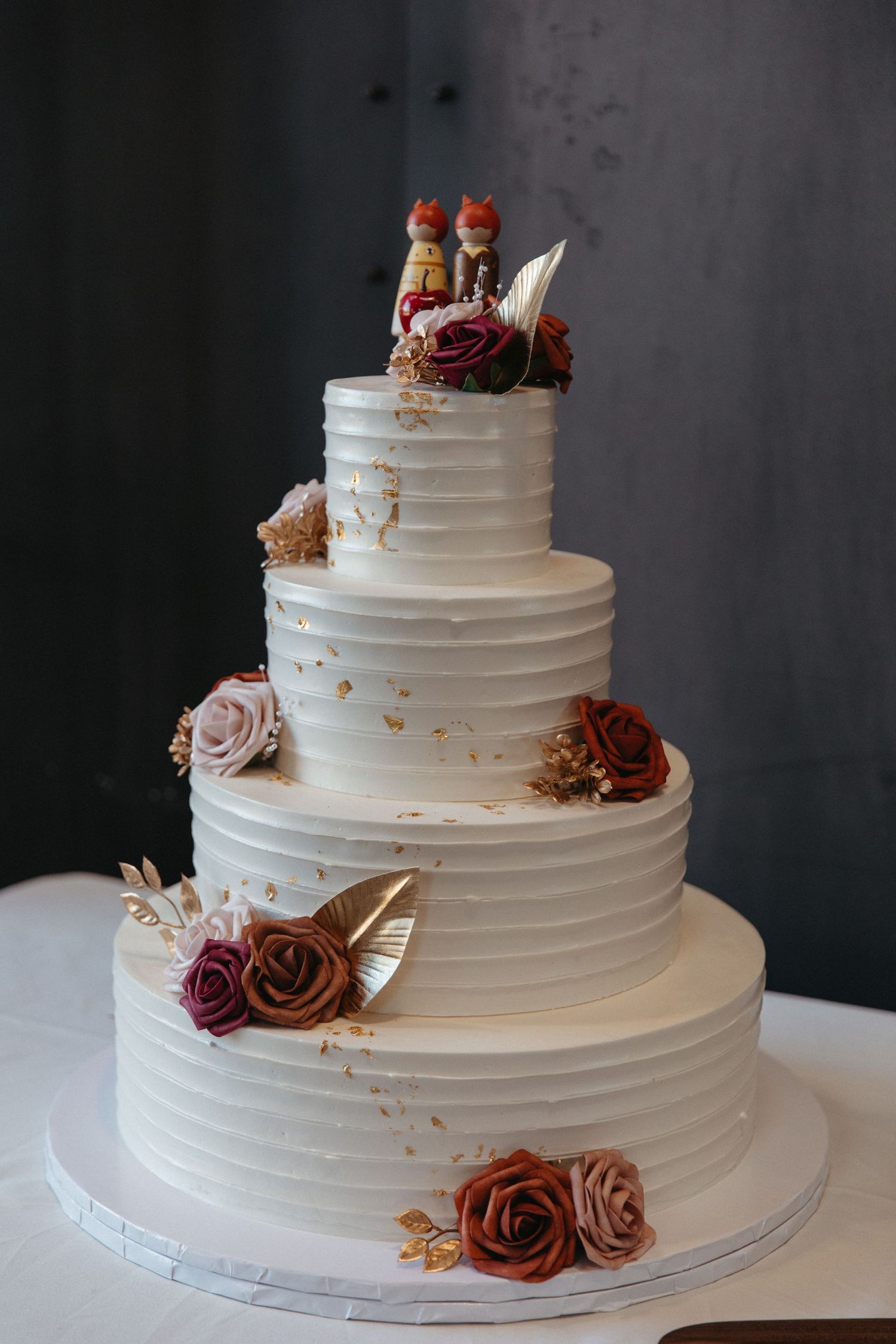 A four-tier white wedding cake decorated with burgundy and dusty rose flowers and gold accents.