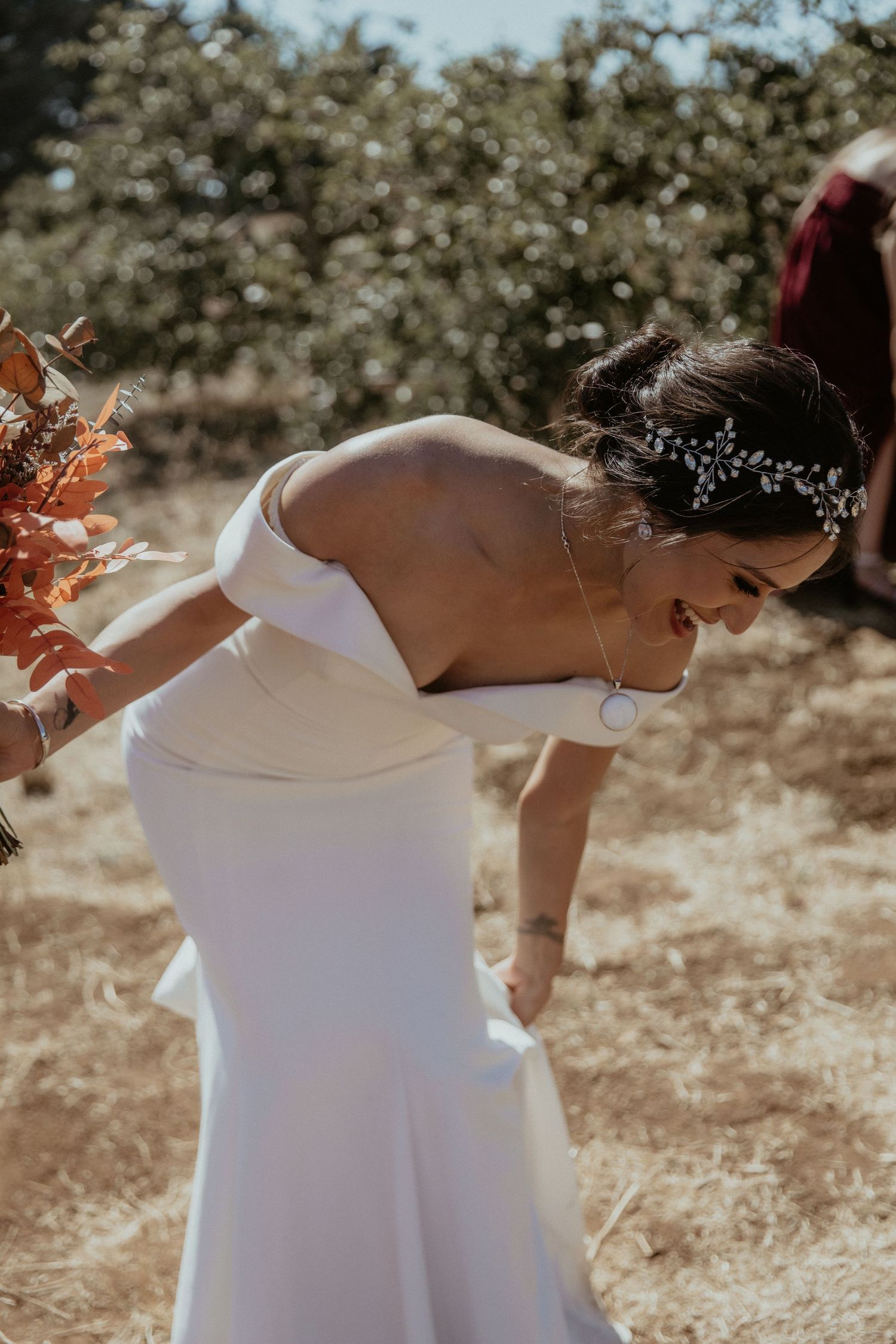A bride in an elegant white off-shoulder dress holds a vibrant autumn-colored bouquet in sunlit outdoor setting.