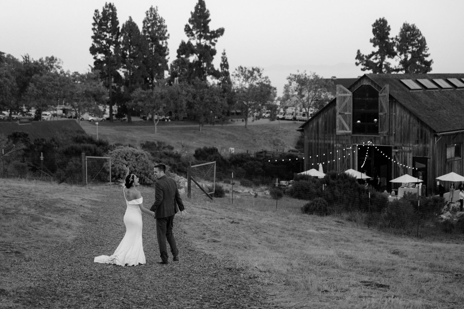 A black and white photo of a couple standing on a hillside near a rustic barn.