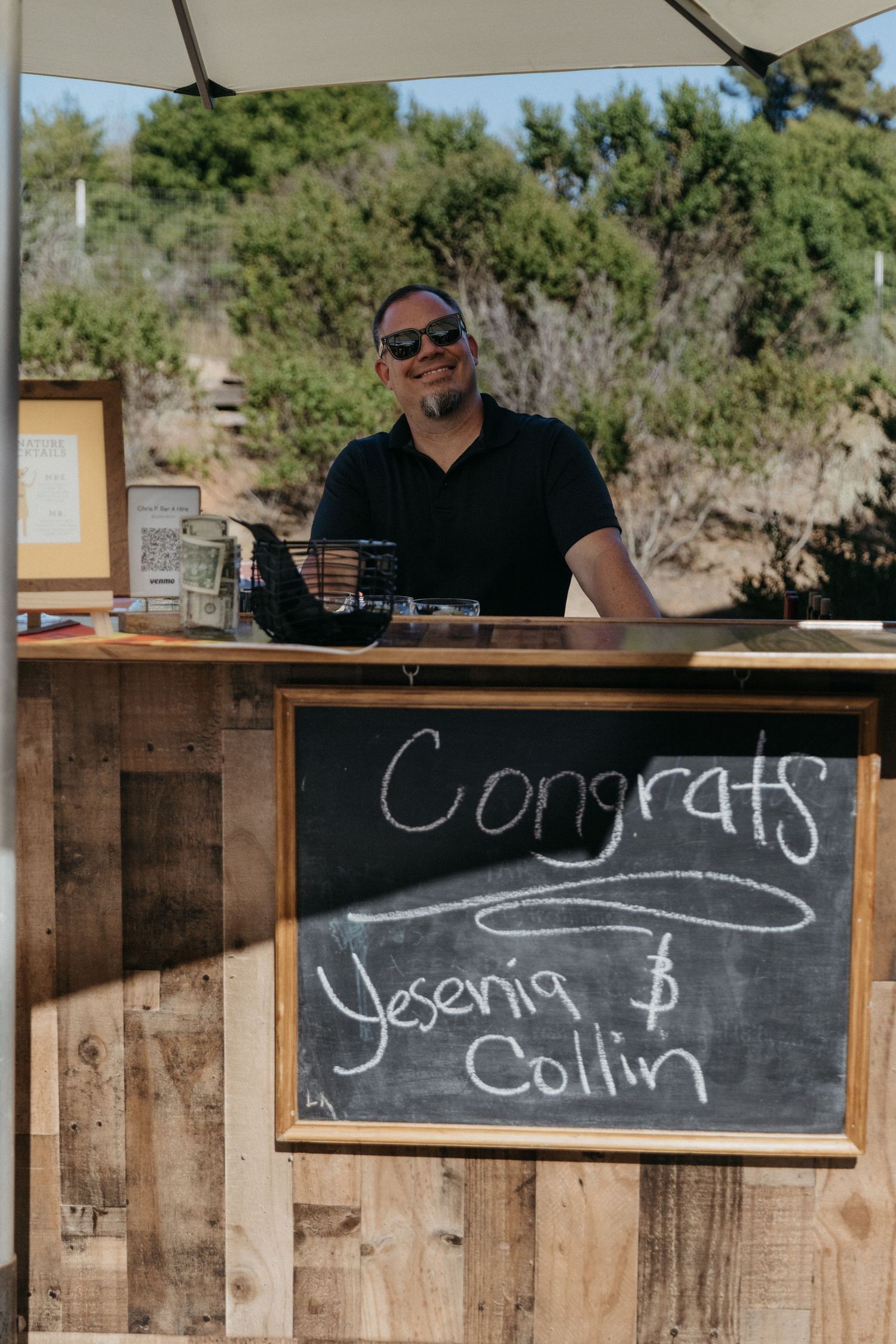 A bartender stands behind a rustic wooden bar with a congratulatory chalkboard sign.