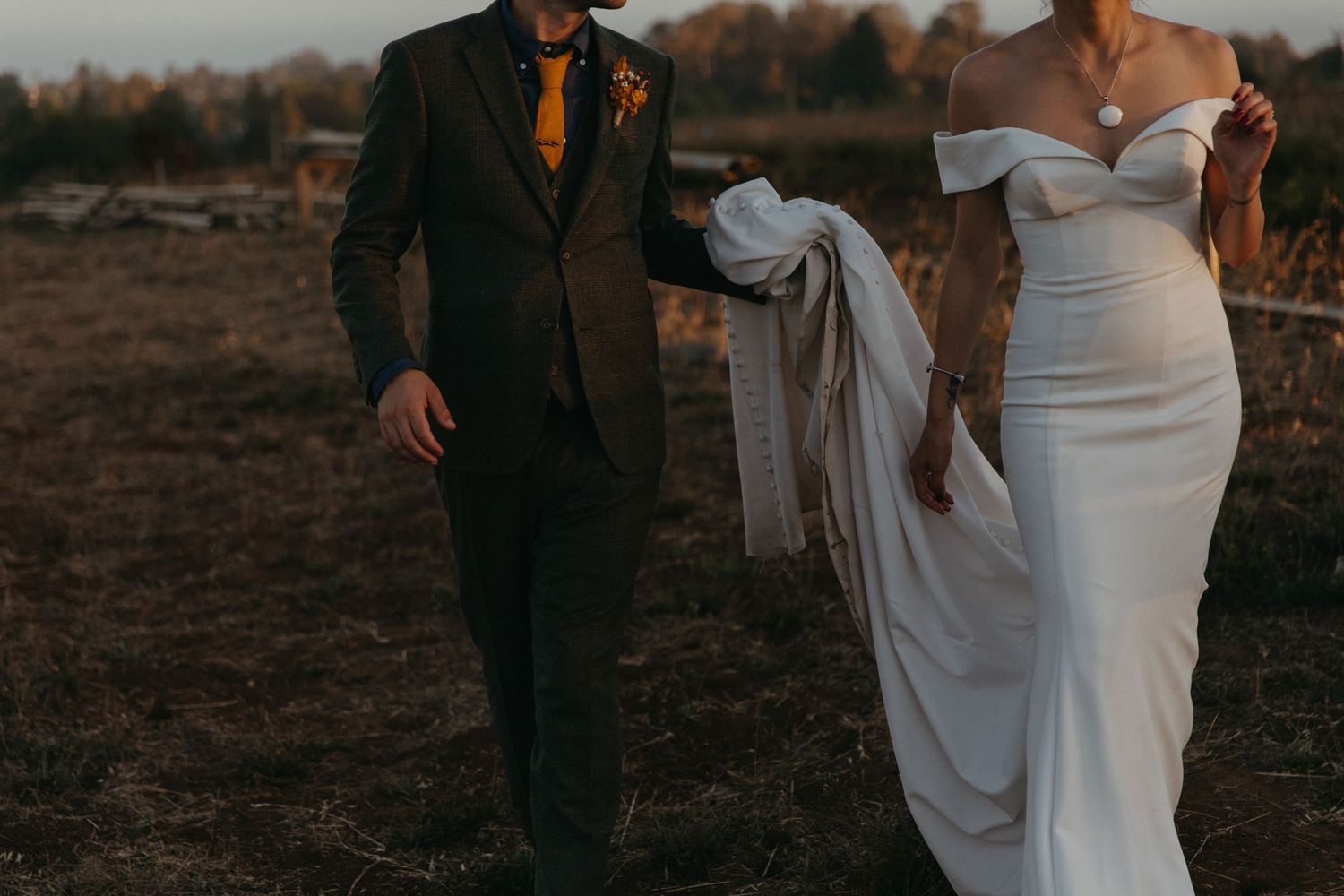 Couple in wedding attire walking through a field at sunset with flowing white dress.