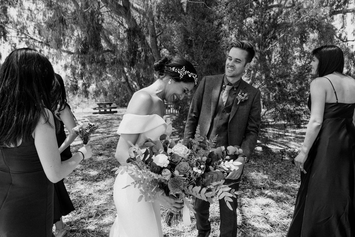 A wedding moment captured in black and white with a bridal bouquet and guests under trees.