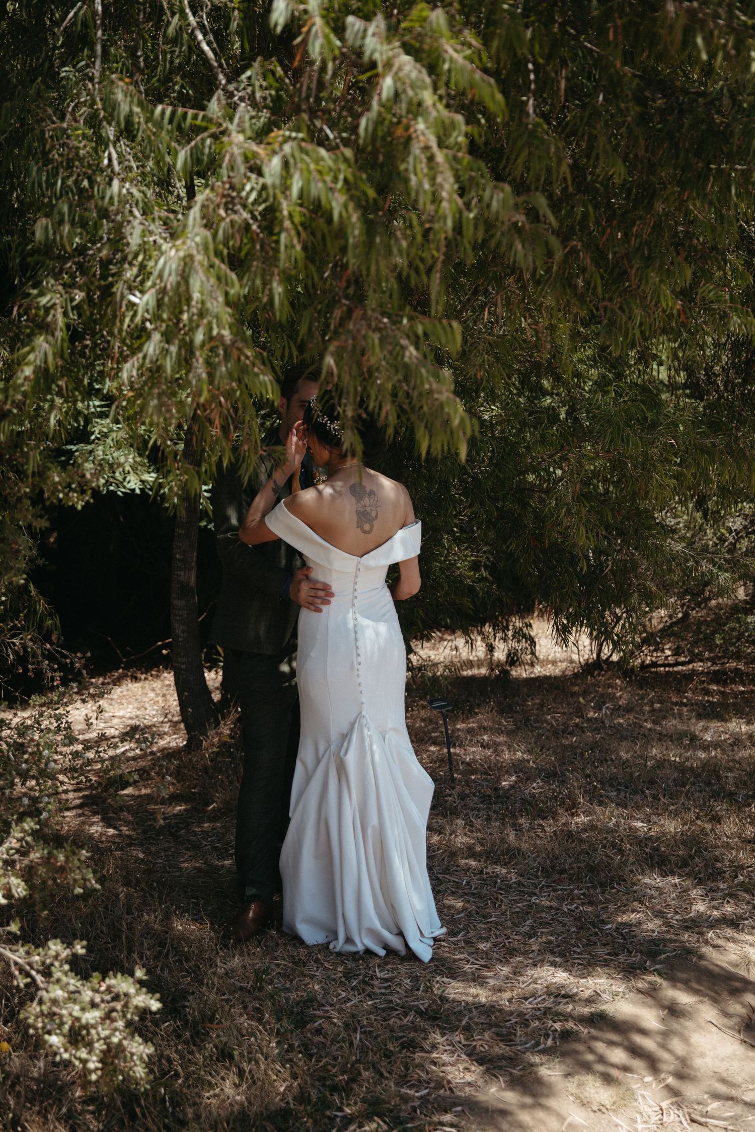 A romantic wedding portrait of a couple embracing under a tree in a sun-dappled outdoor setting.