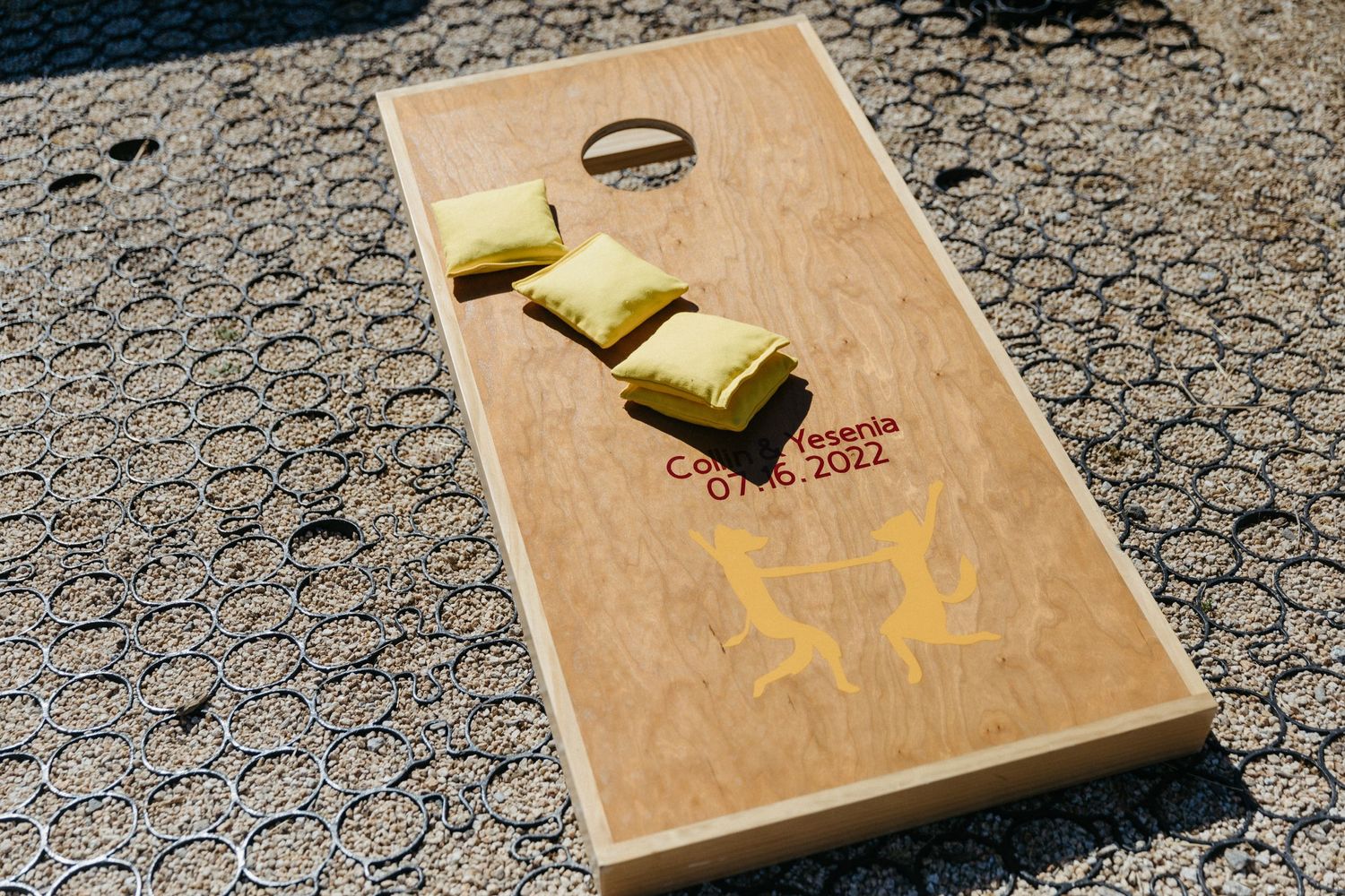 A wooden cornhole game board with yellow beanbags and a ring displayed on top.