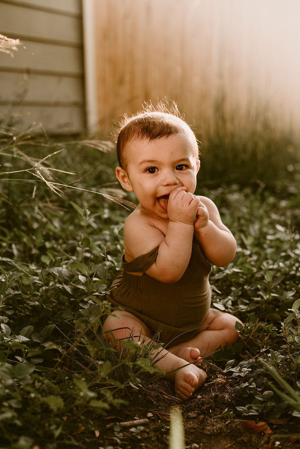 Baby in brown romper playing in garden with hands near mouth during sunset.
