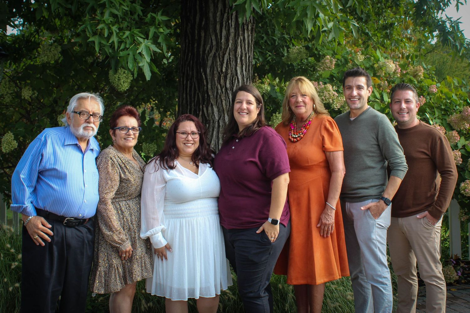Family gathering photo with seven people posed in front of lush garden scenery.