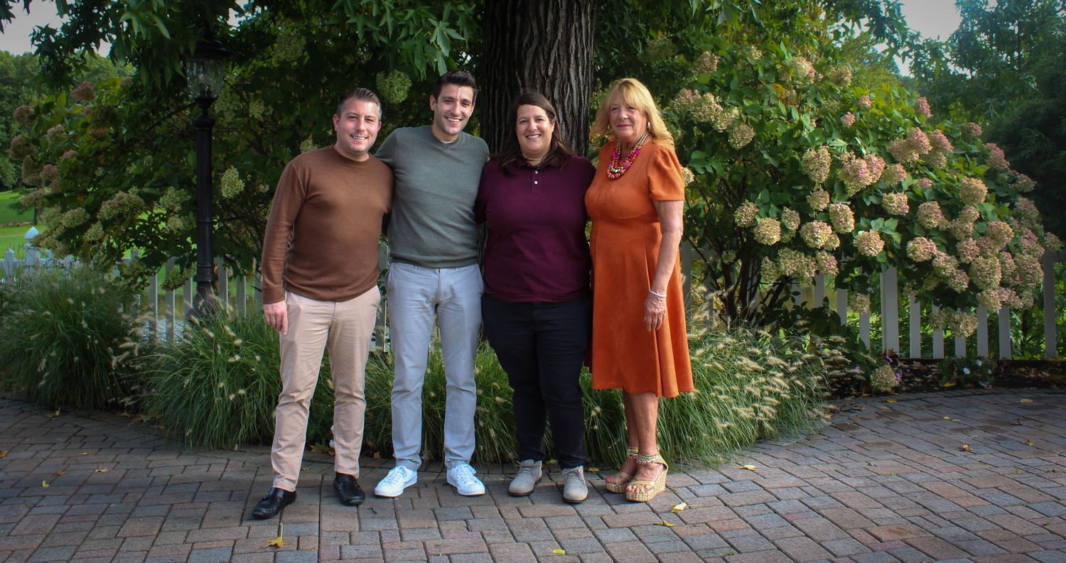 A group of four people stand together on a brick path in front of lush garden greenery and hydrangea flowers.