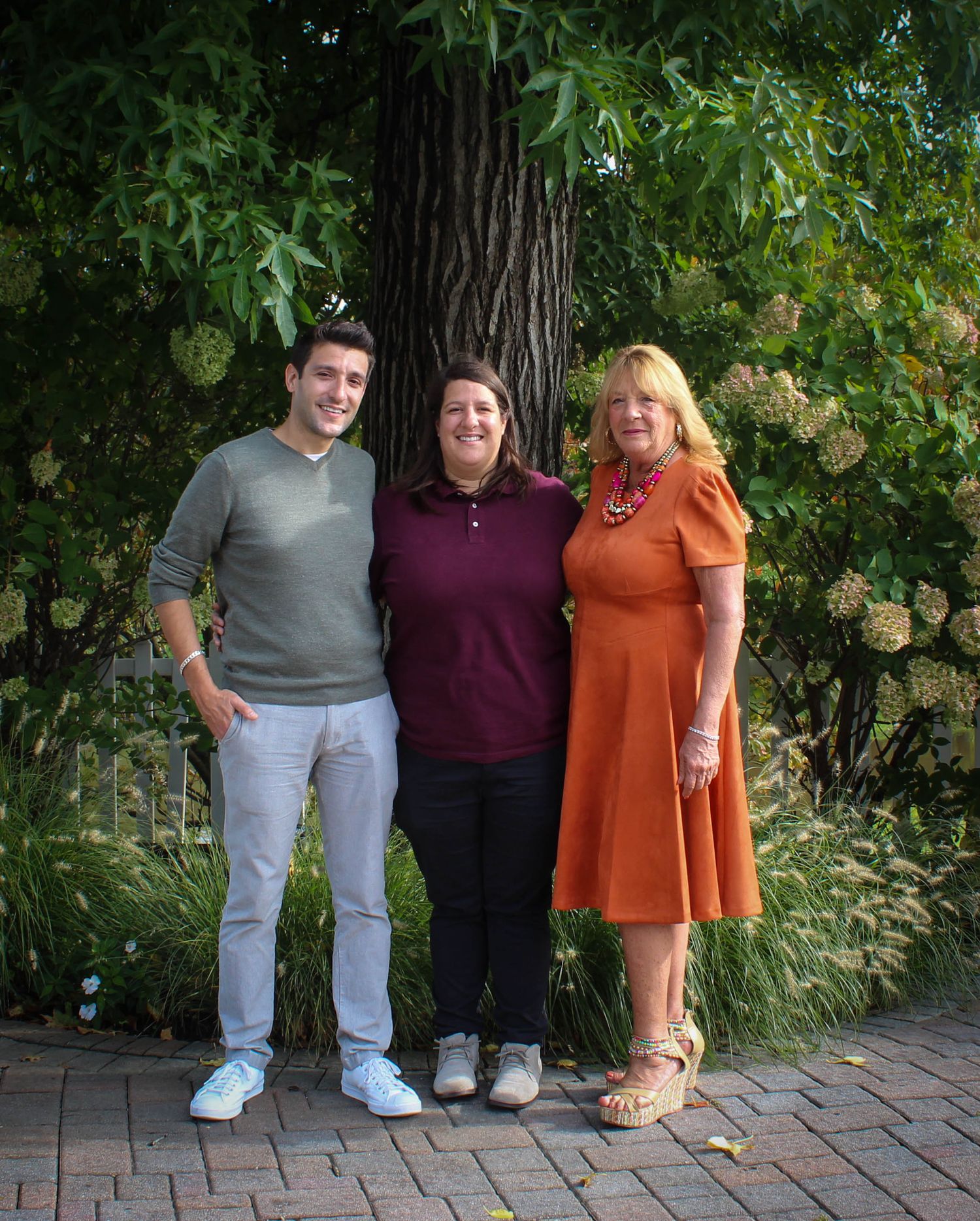 Three people stand together on a brick path with a large tree trunk and flowering shrubs in the background.