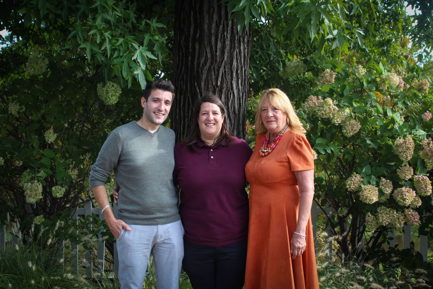 Three people pose for a casual outdoor photo with flowering shrubs and trees in the background.