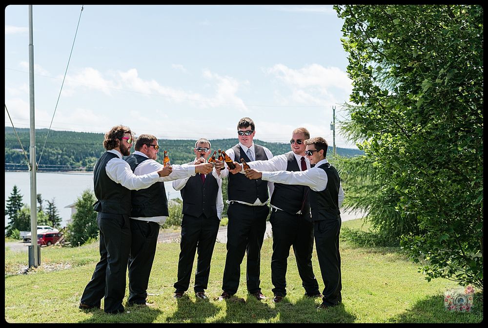 A group of groomsmen in formal attire gather for a photo by a lake.