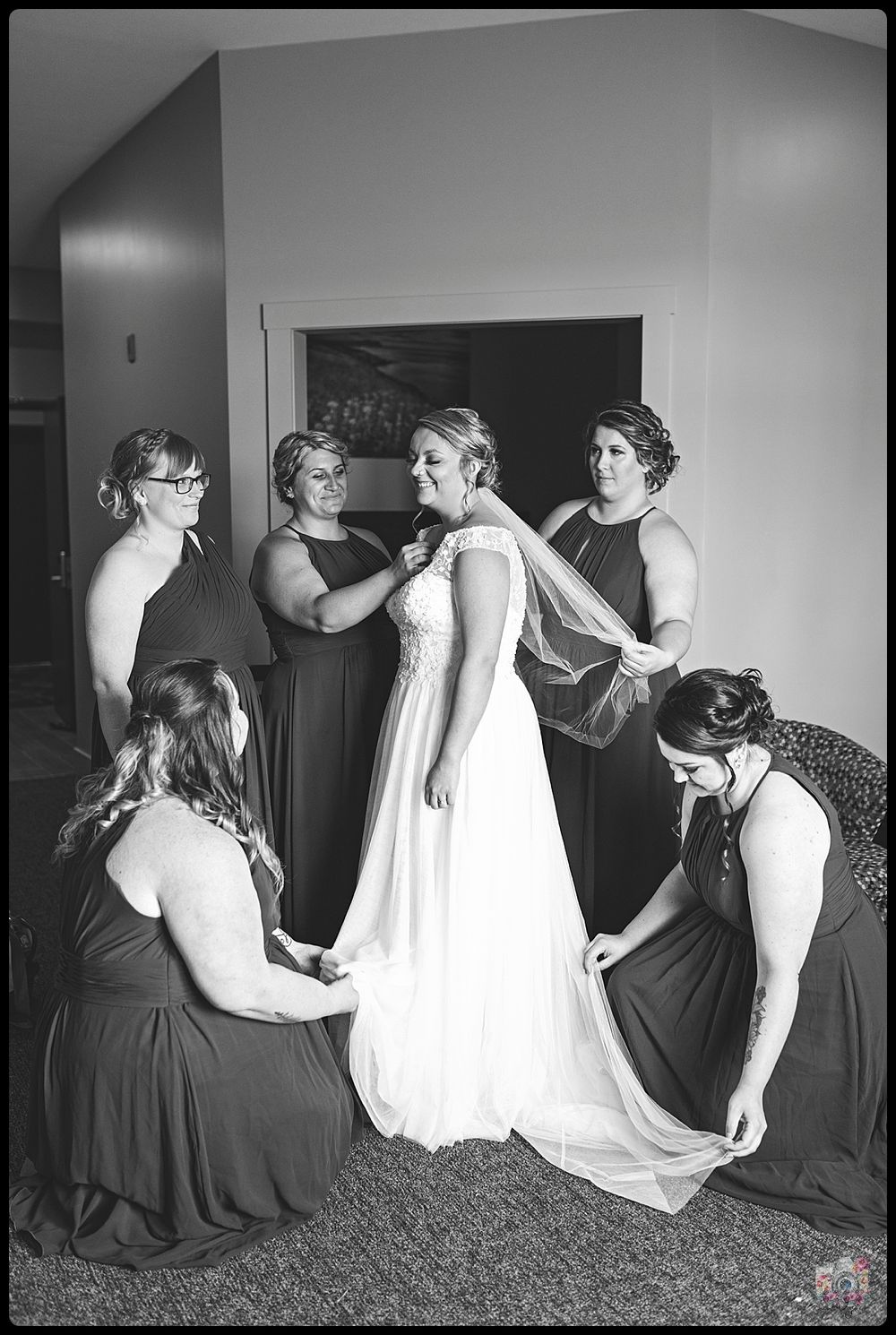 Bridesmaids in dark dresses help adjust a white wedding gown during pre-ceremony preparations.