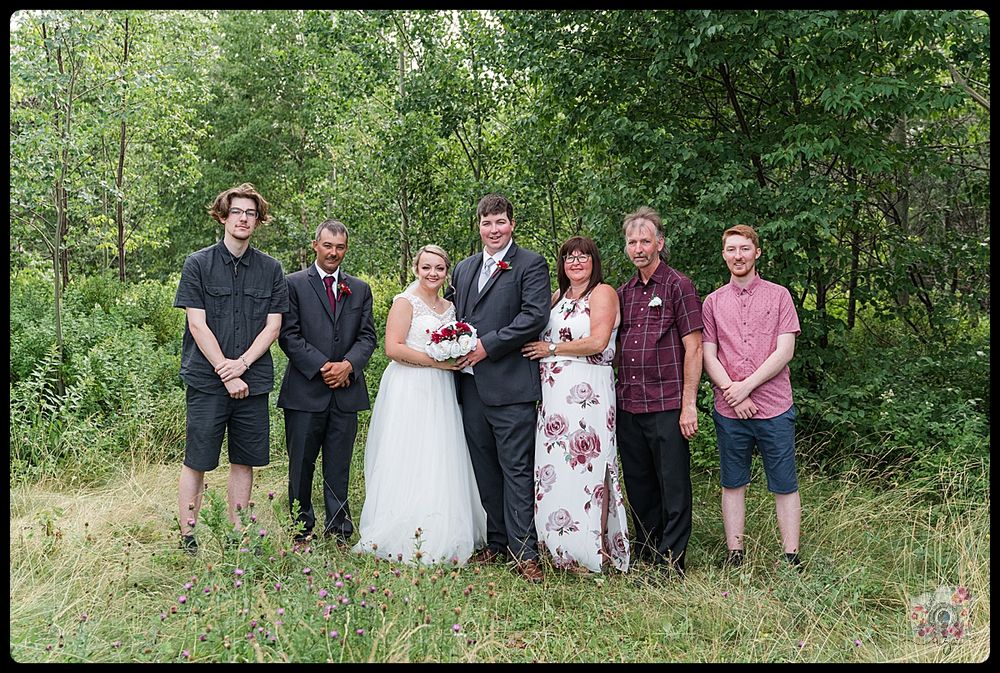 Wedding party members gather for a casual group photo in a wooded outdoor setting.